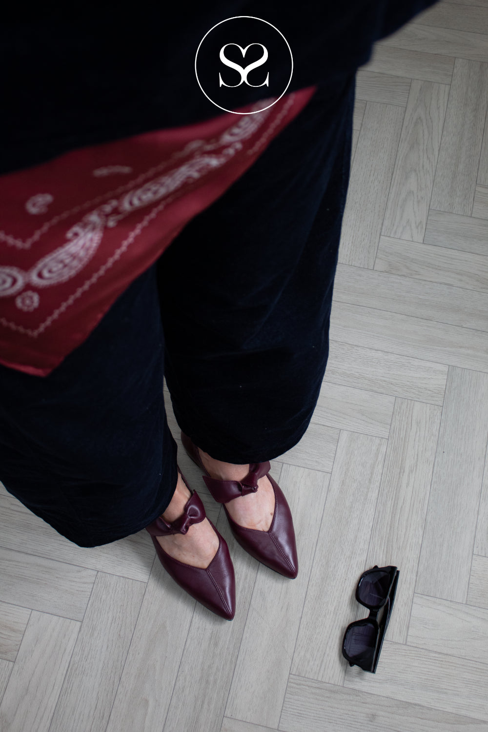A person wearing dark navy velvet pants and burgundy pointed-toe Mary Jane flats with bow details, standing on a light gray herringbone wood floor. A red paisley bandana is draped over the leg, and black sunglasses are on the floor nearby.