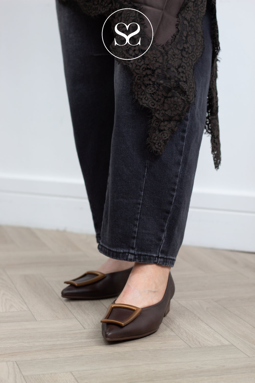A person wearing dark brown pointed-toe flats with gold-tone square buckles, paired with dark jeans and a black lace-trimmed garment, standing on a light wood floor against a white wall.