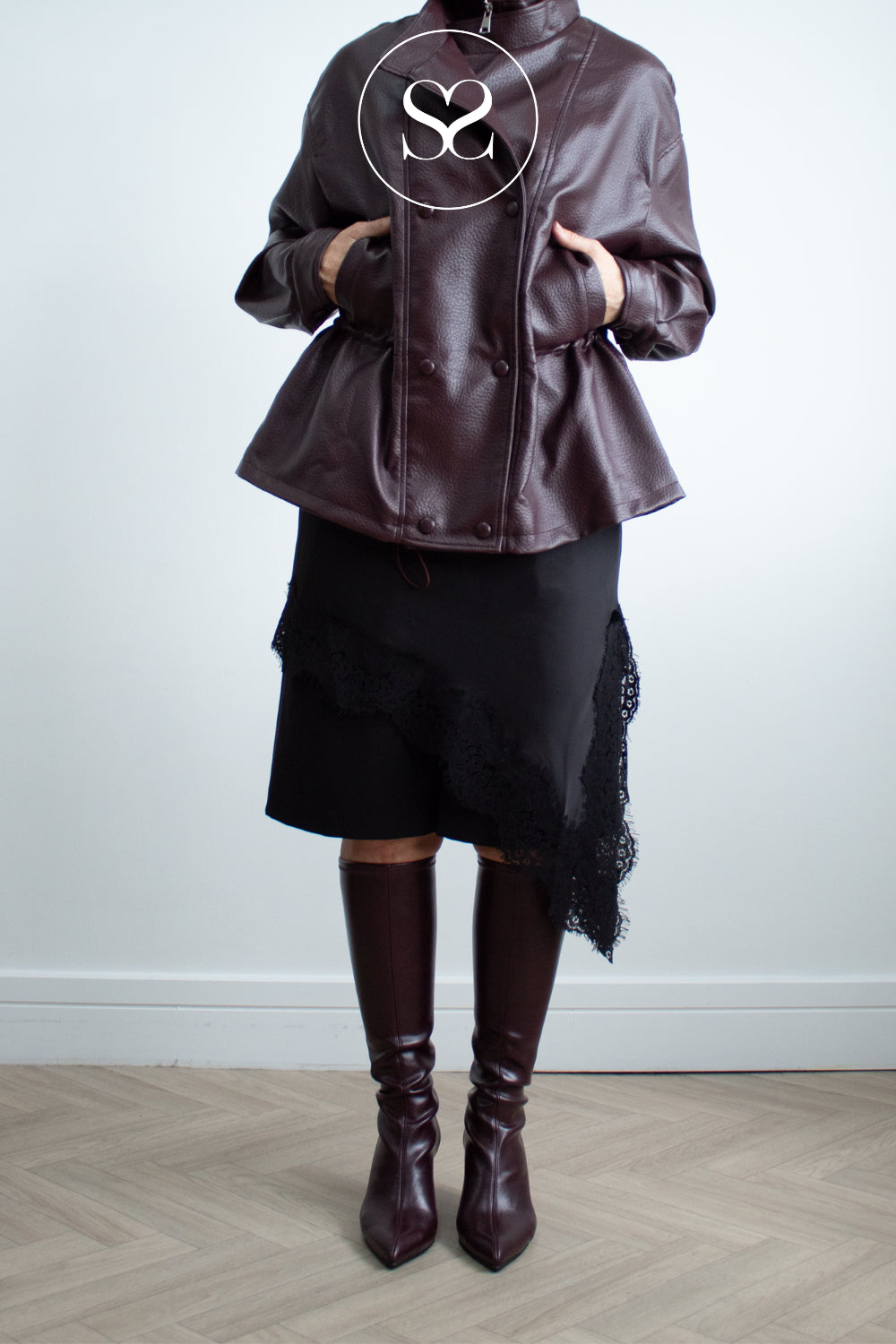 A model wearing a dark brown faux-leather peplum jacket, a black asymmetrical skirt with lace trim, and matching dark burgundy Miss ELastic pointed-toe boots, standing against a light gray wall and wood herringbone floor.