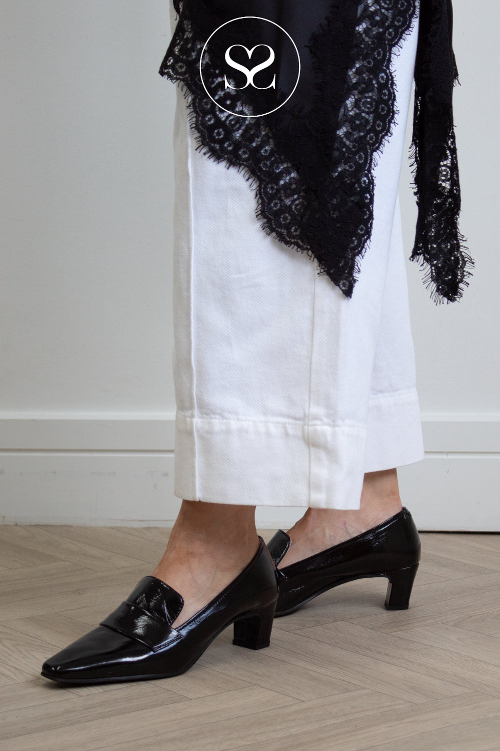 A person wearing black patent leather loafers with block heels from Unisa, styled with white cropped trousers and a black lace shawl, standing on a light wood floor.