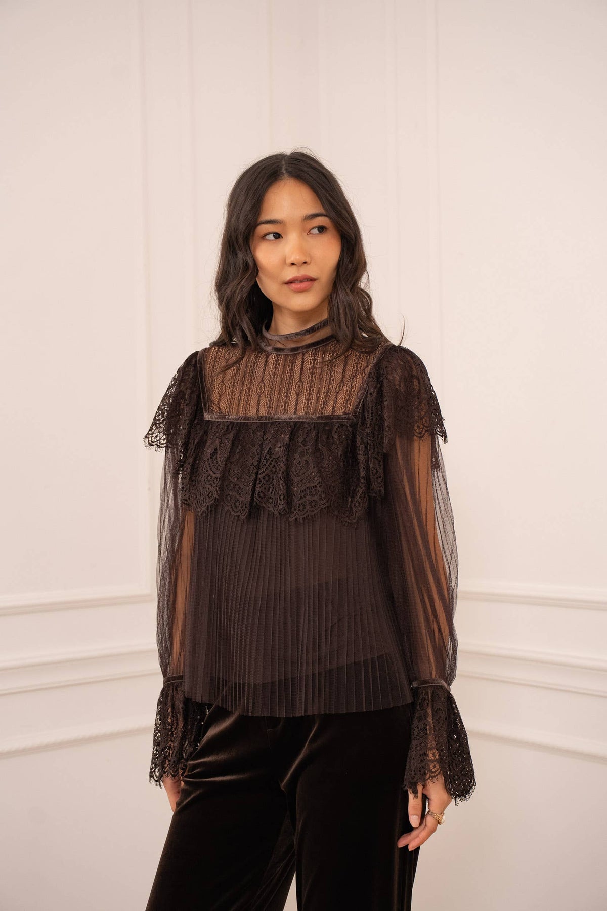 Woman wearing a dark brown lace-trimmed blouse against a white wall