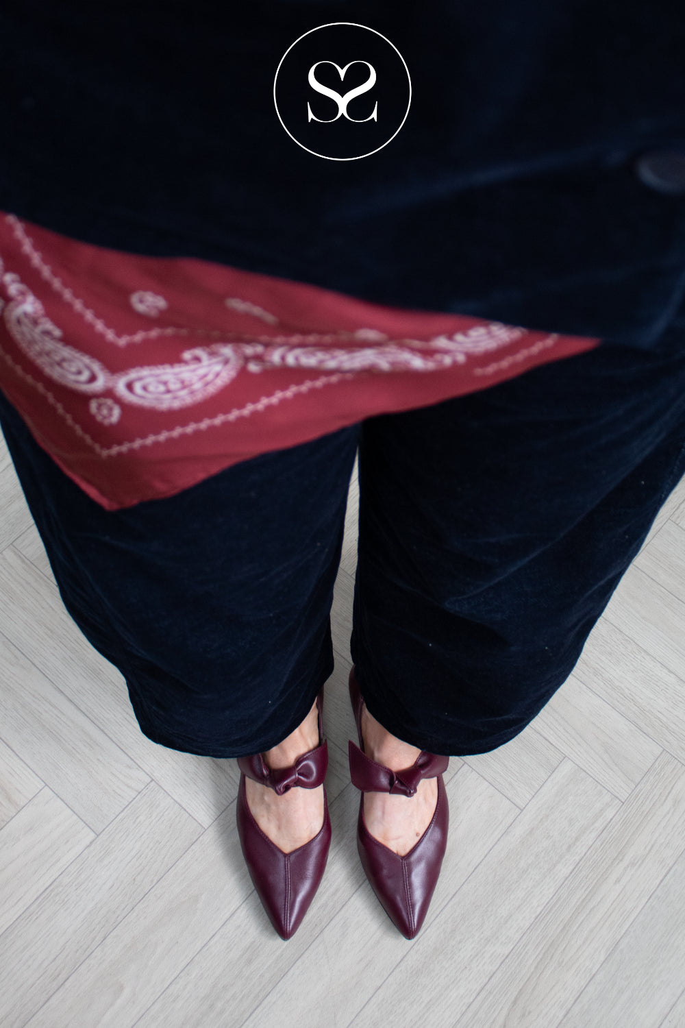 A person wearing dark navy velvet pants and burgundy pointed-toe flats with bow details, standing on a light wood floor. A red paisley bandana is draped over the waist, and a circular 'SS' logo watermark is visible at the top.