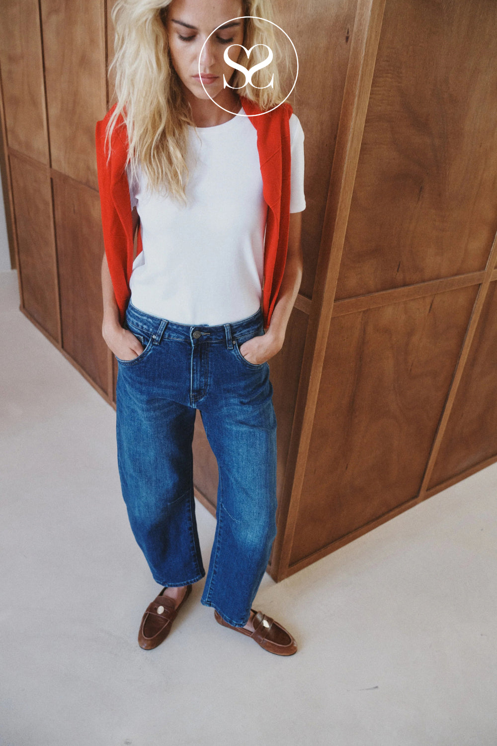 A woman wearing blue barrel ankle length jeans, a white t-shirt, a red cardigan, and brown loafers, standing in front of a wooden paneled wall.