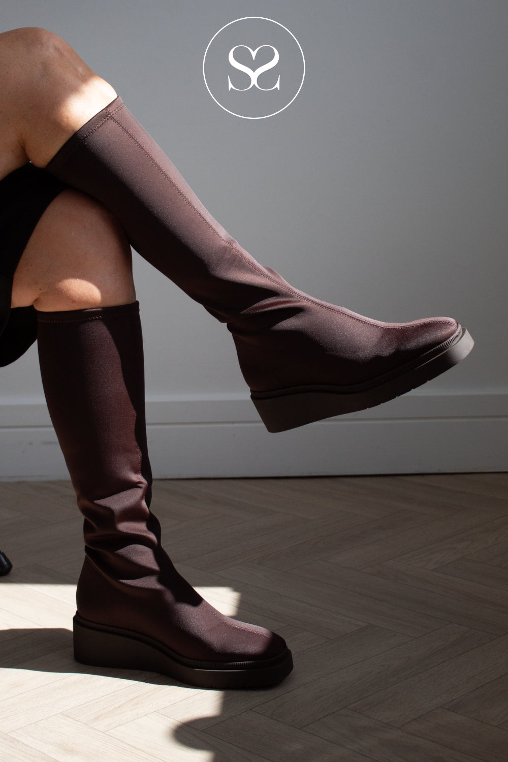 A woman wearing Miss Elastic dark brown knee-high platform boots with a draped, elasticized fabric upper, shown in a styled indoor setting with a gray wall and wooden floor. in Sheneil Shoes Galway Ireland