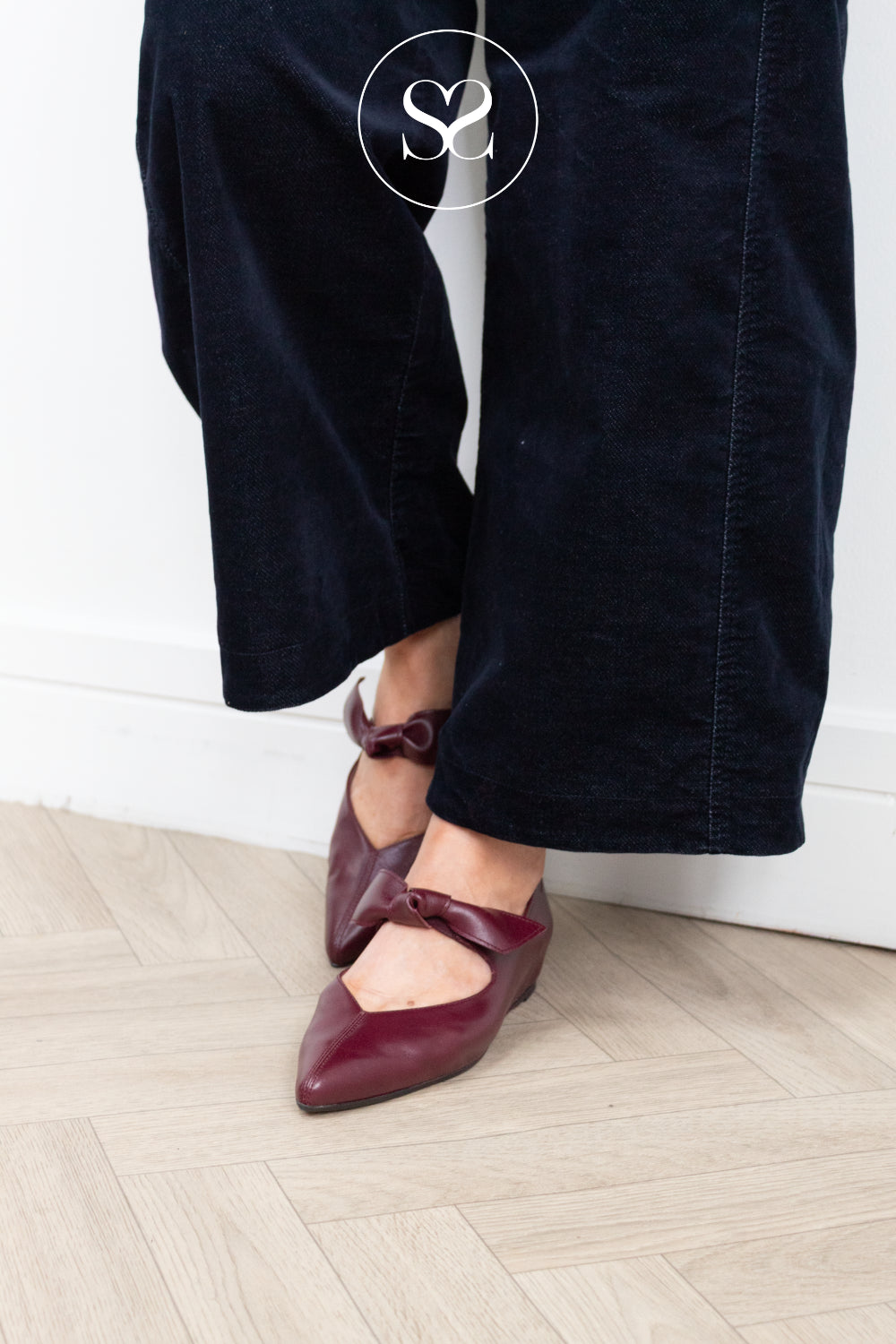 A person wearing dark navy wide-leg corduroy trousers and burgundy pointed-toe flats with ankle bows, standing on a light wood herringbone floor against a white wall.
