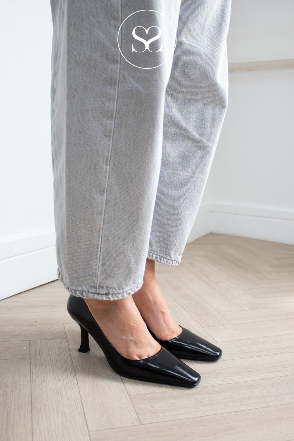 A woman wearing light gray wide-leg jeans and black high shine pointed-toe kitten heel pumps, standing on a light wood floor against a white wall. shoes are lodi pauline - available in Ireland