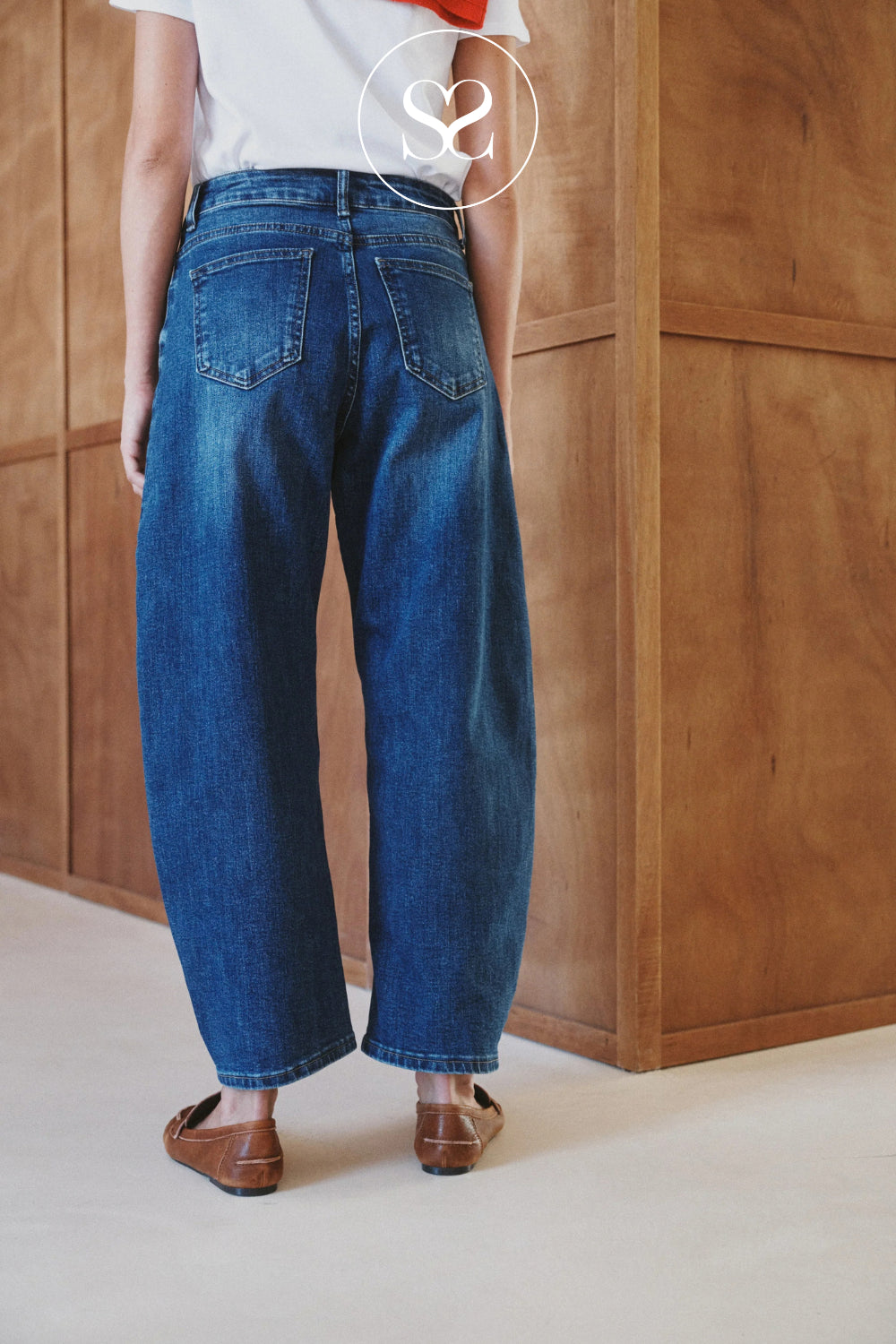 A person shown from the back wearing loose-fit blue denim barel jeans, a white top, and brown loafers in an indoor setting with a wooden wall panel background.