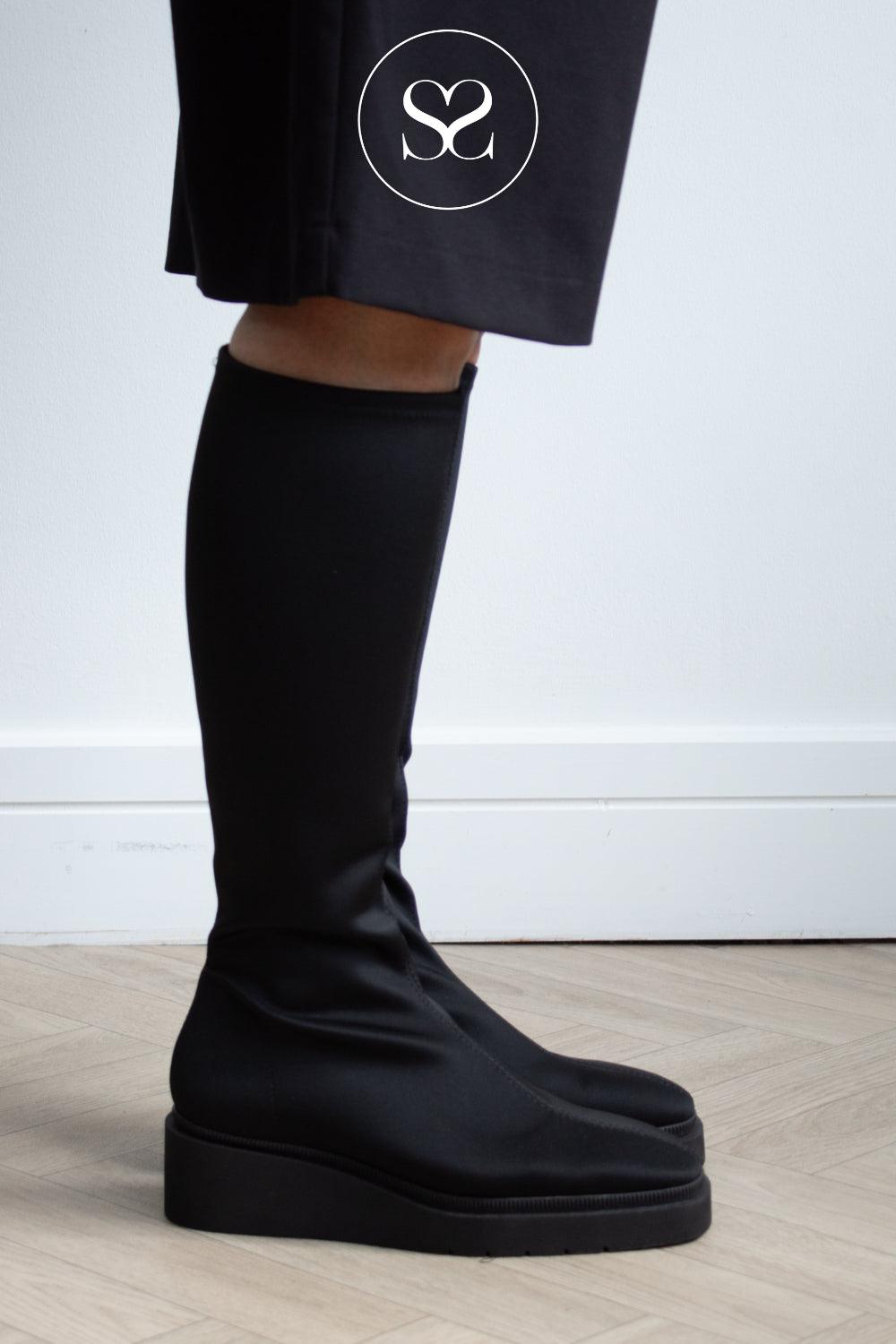 A close-up of a woman's lower legs wearing Miss Elastic black knee-high slip-on boots with chunky platform soles, paired with black trousers featuring a white circular Sheneil SHoes logo.