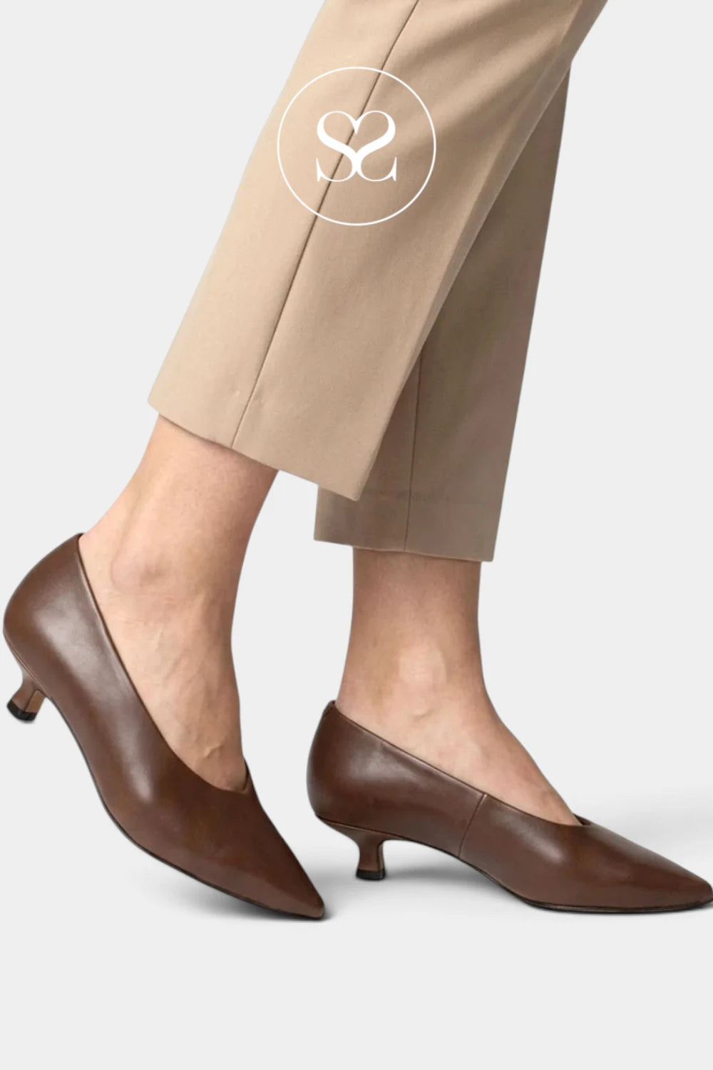 A close-up of a person's legs wearing brown pointed-toe kitten heel pumps with beige trousers, shown against a light gray studio background. A white circular 'SC' logo is overlaid near the top.