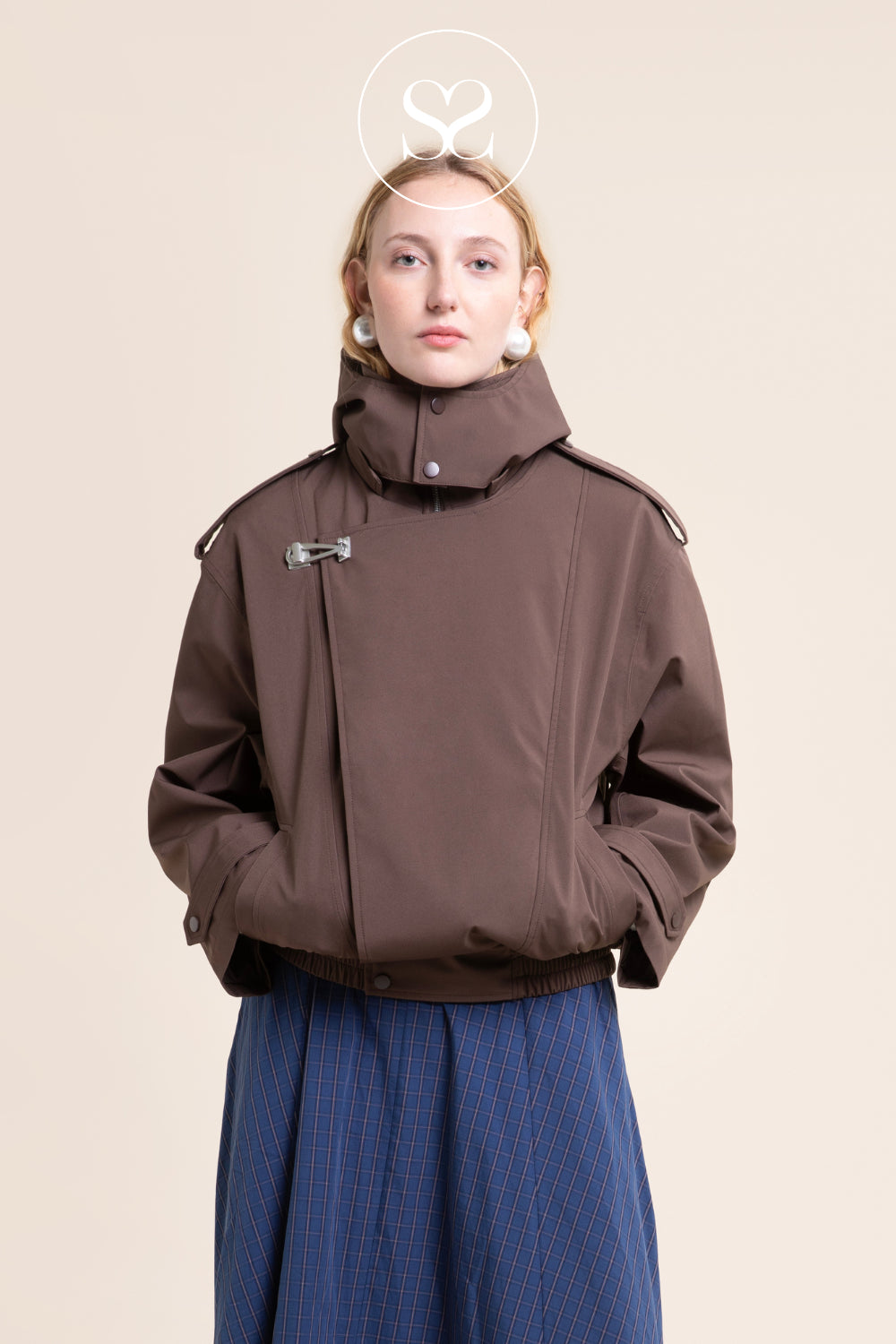 A model wearing a brown asymmetrical high-collar jacket with a unique silver-toned snap closure from Flotte and a blue striped skirt, set against a solid beige background.
