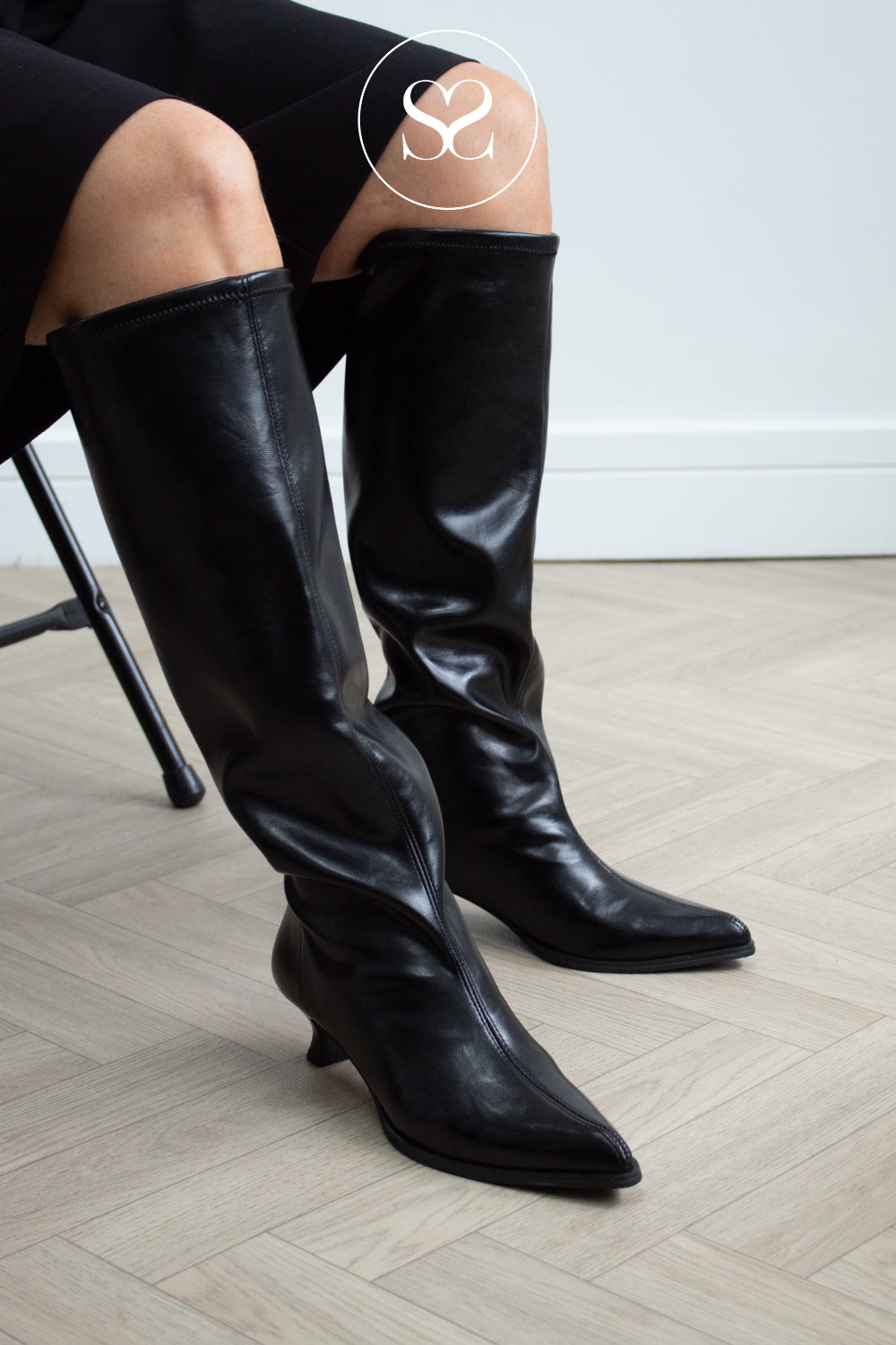 A pair of black leather look knee-high boots with pointed toes and low kitten heels, worn by a model sitting on a chair on a light wood herringbone floor. 
