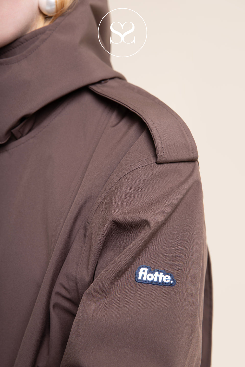 Close-up of a person wearing a dark brown hooded jacket, showing the shoulder and upper arm with a blue and white 'flotte.' logo patch on the sleeve, against a light beige background in Ireland