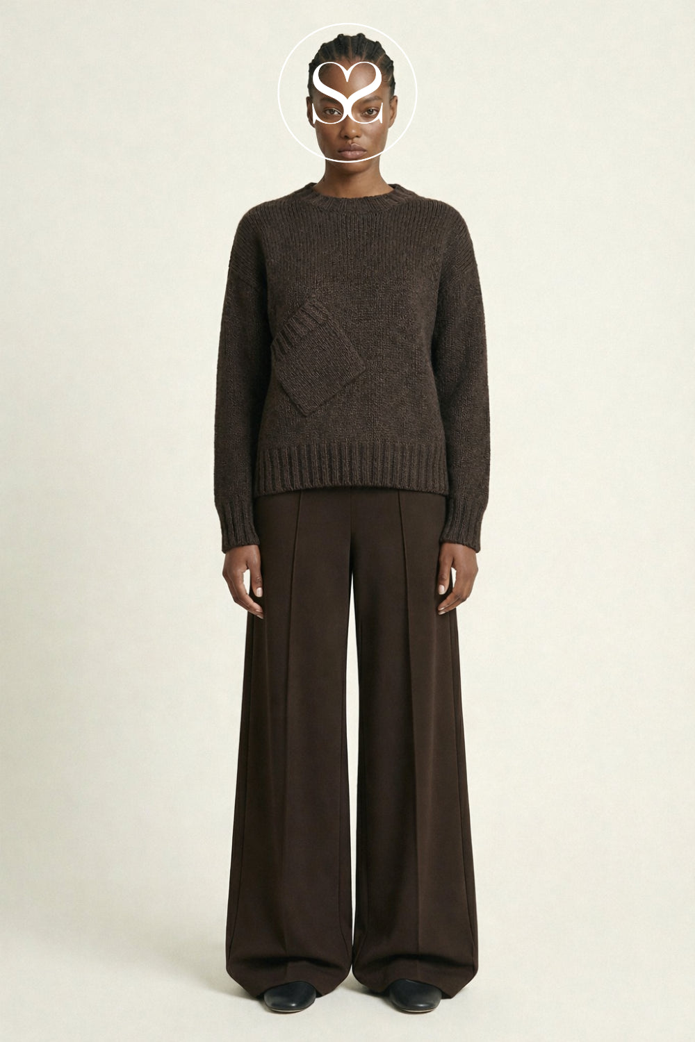 A model wearing a dark brown knit sweater with a textured diamond pattern on the chest and matching dark brown wide-leg trousers, standing against a light beige studio background.