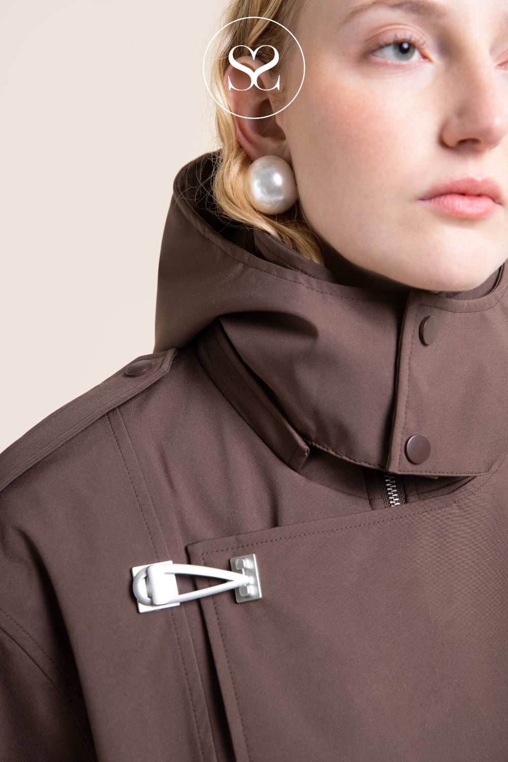 Close-up of a model wearing a brown hooded jacket with a silver metal toggle closure on the chest, a pearl earring, and a circular 'Sheneil Shoes Ireland' logo watermark near the top.