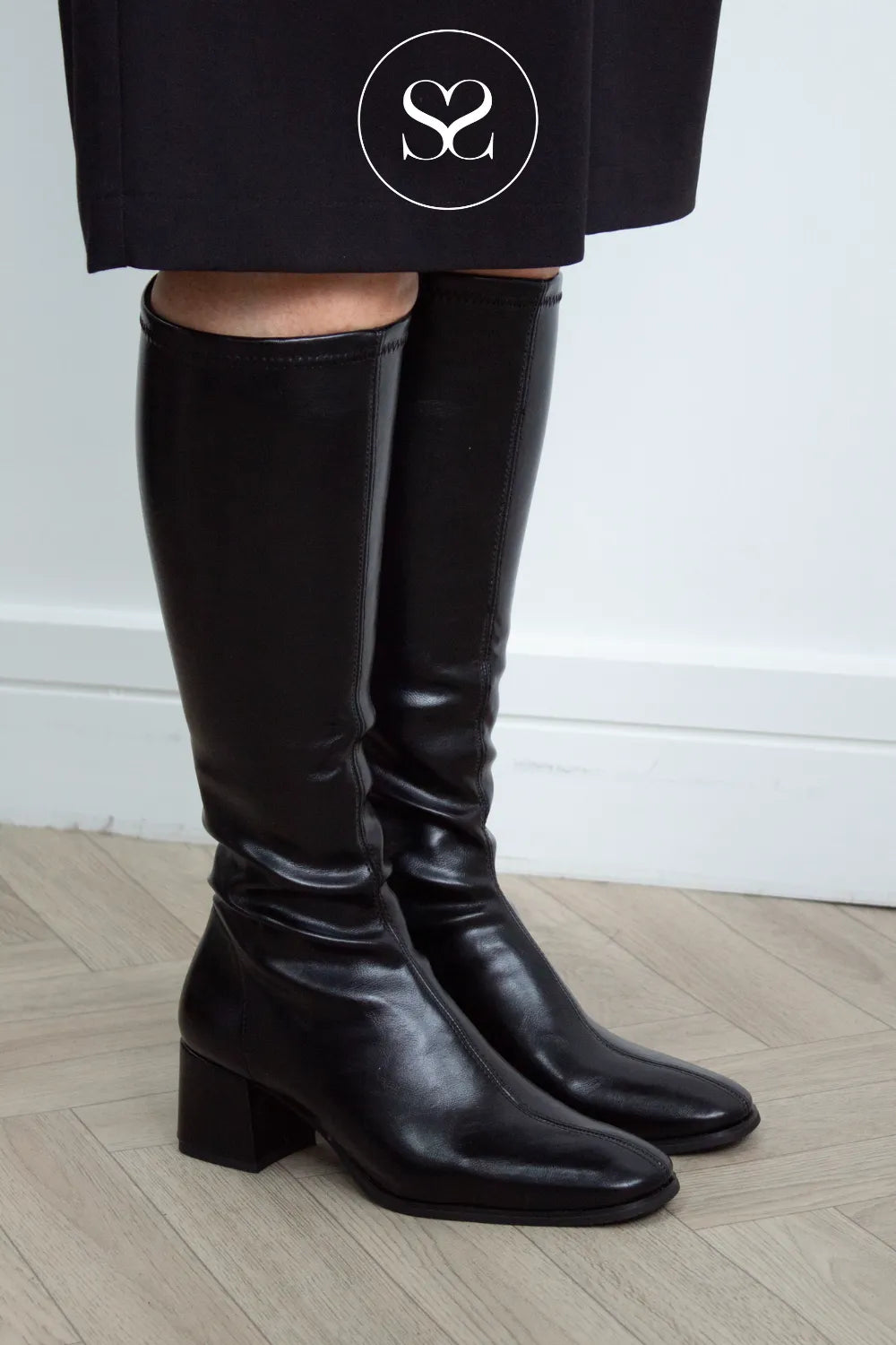 A pair of black leather look knee-high sock boots with a block heel, shown on a model wearing a black skirt. The background is a clean white wall and light wood floor. Miss elastic boots imaged by sheneil shoes Galway Ireland