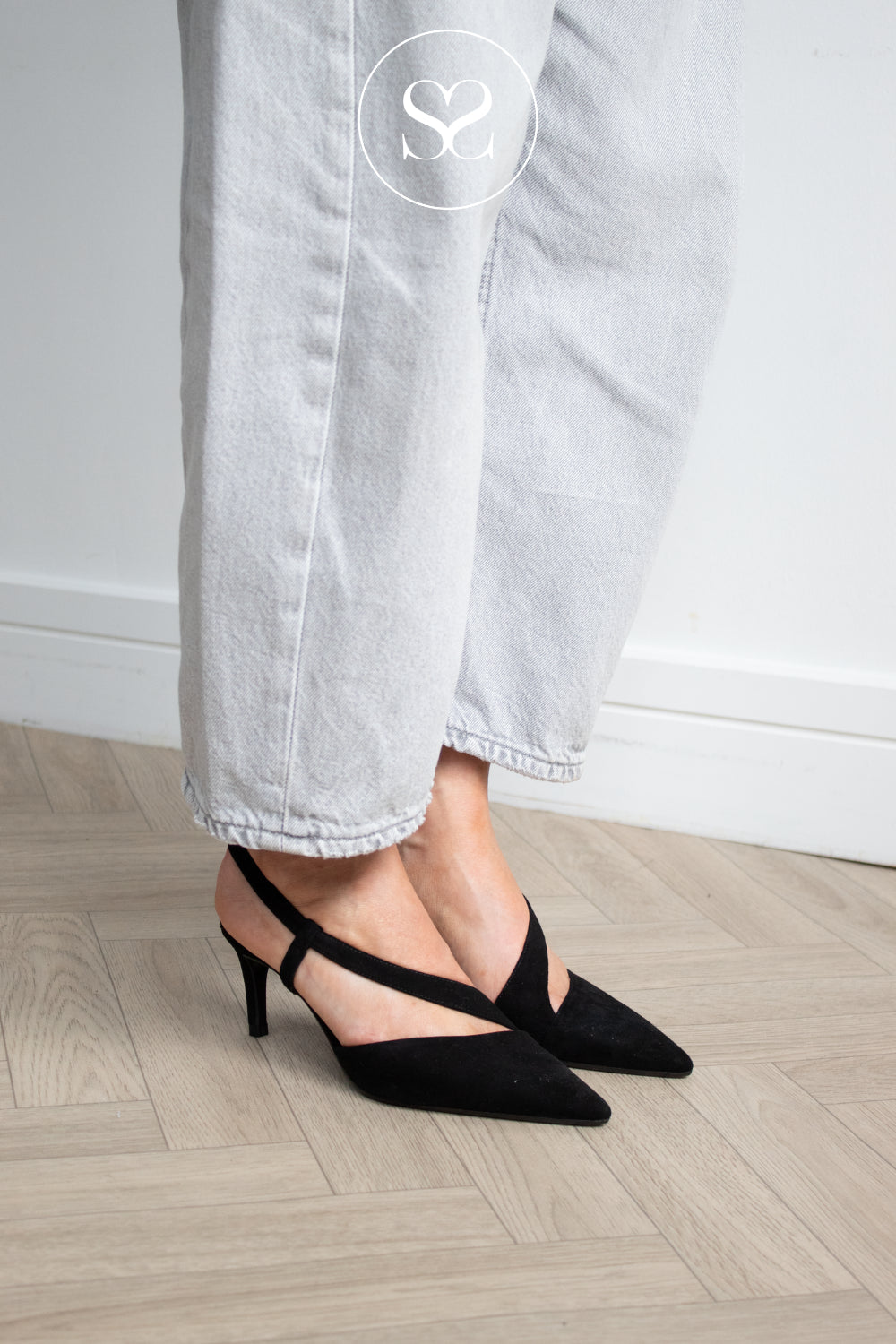 Black Suede slingback pointed-toe heels with crisscrossing front straps, shown on a model wearing light gray jeans on a light wood herringbone floor. Heels are lodi Naltom style