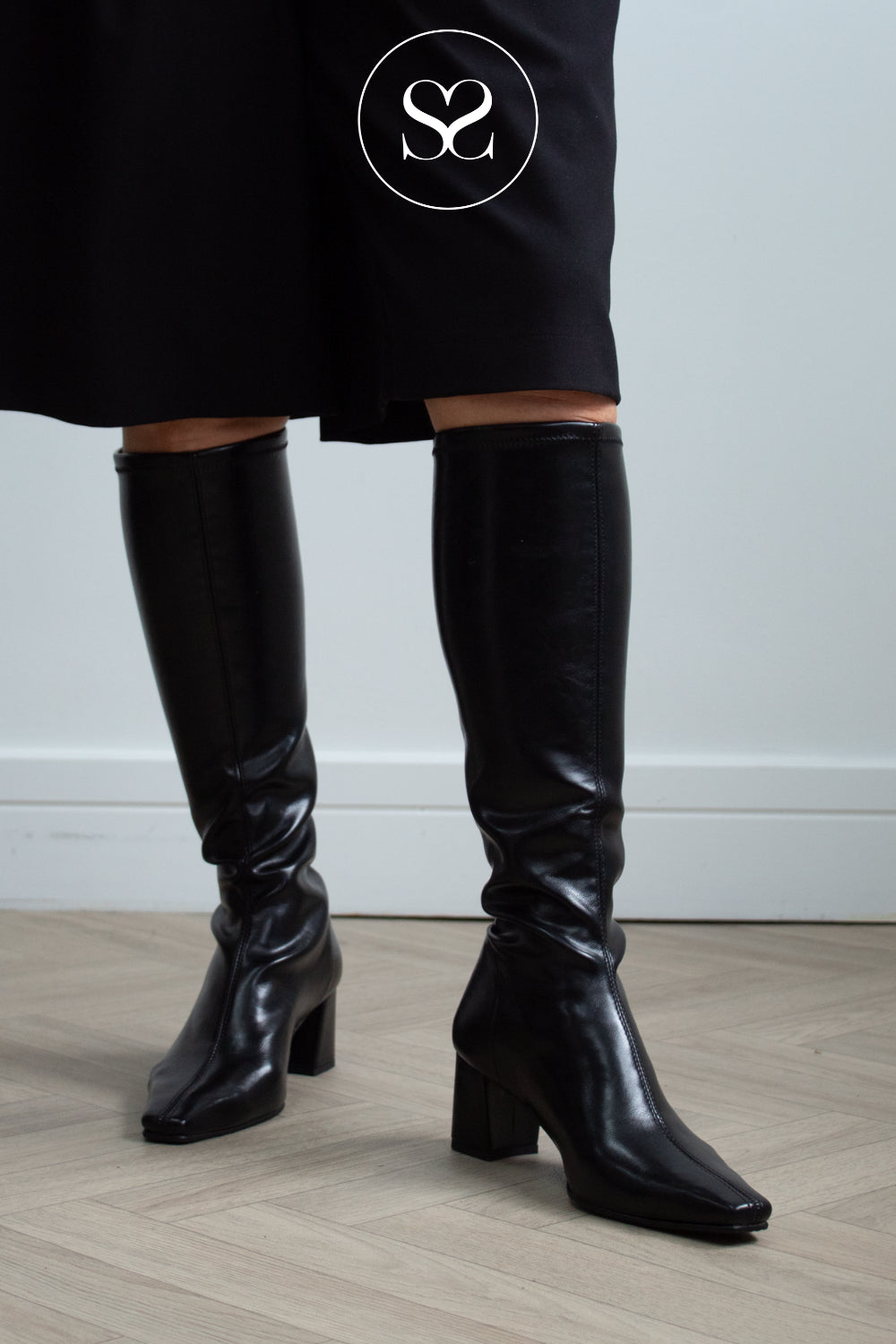 A person wearing Miss Elastic shiny black knee-high boots with block heels and pointed toes, paired with a black skirt. The photo is cropped from the upper skirt hem down to the feet, set against a light gray wall and pale wood floor.