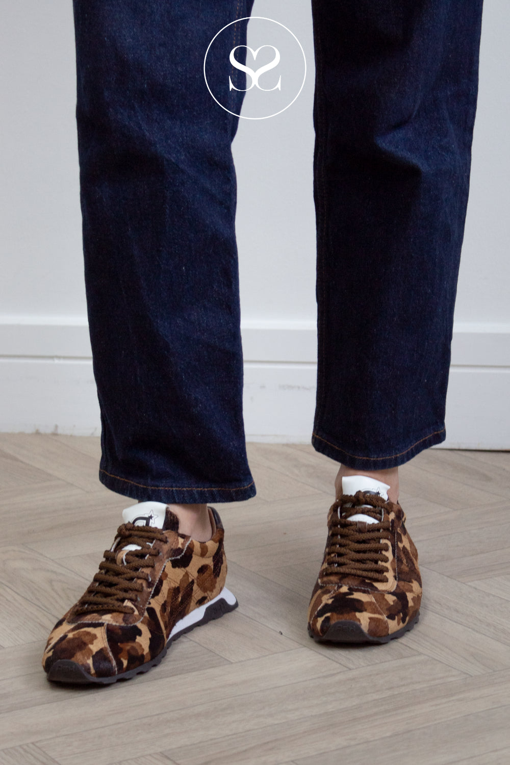 A woman wearing Ash dark brown and tan leopard-print sneakers with dark blue jeans, standing on a light wood floor against a white wall.
