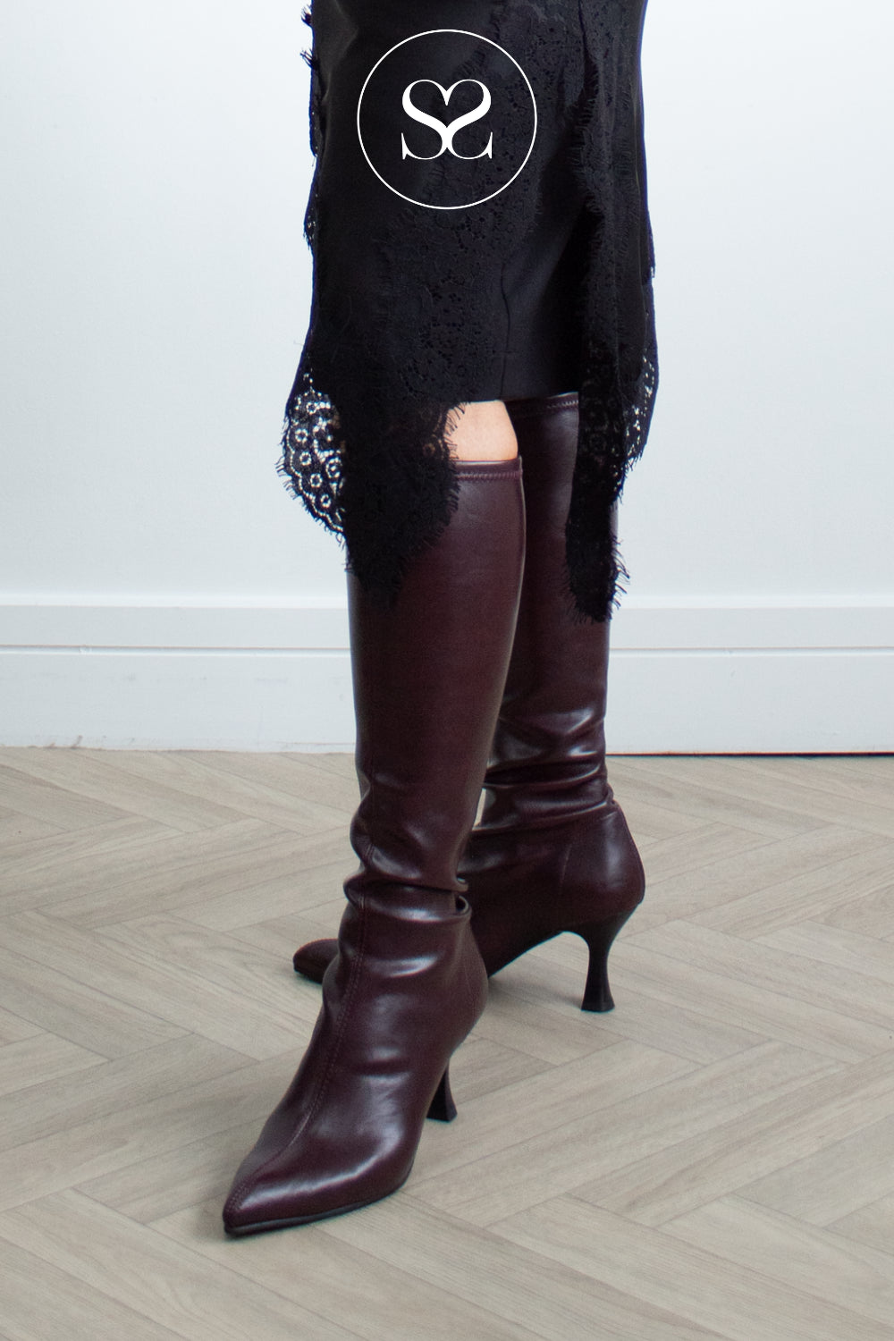 A pair of dark burgundy leather knee-high heeled sock boots with pointed toe worn with a black lace-trim top, shown from the knees down on a light wood herringbone floor.