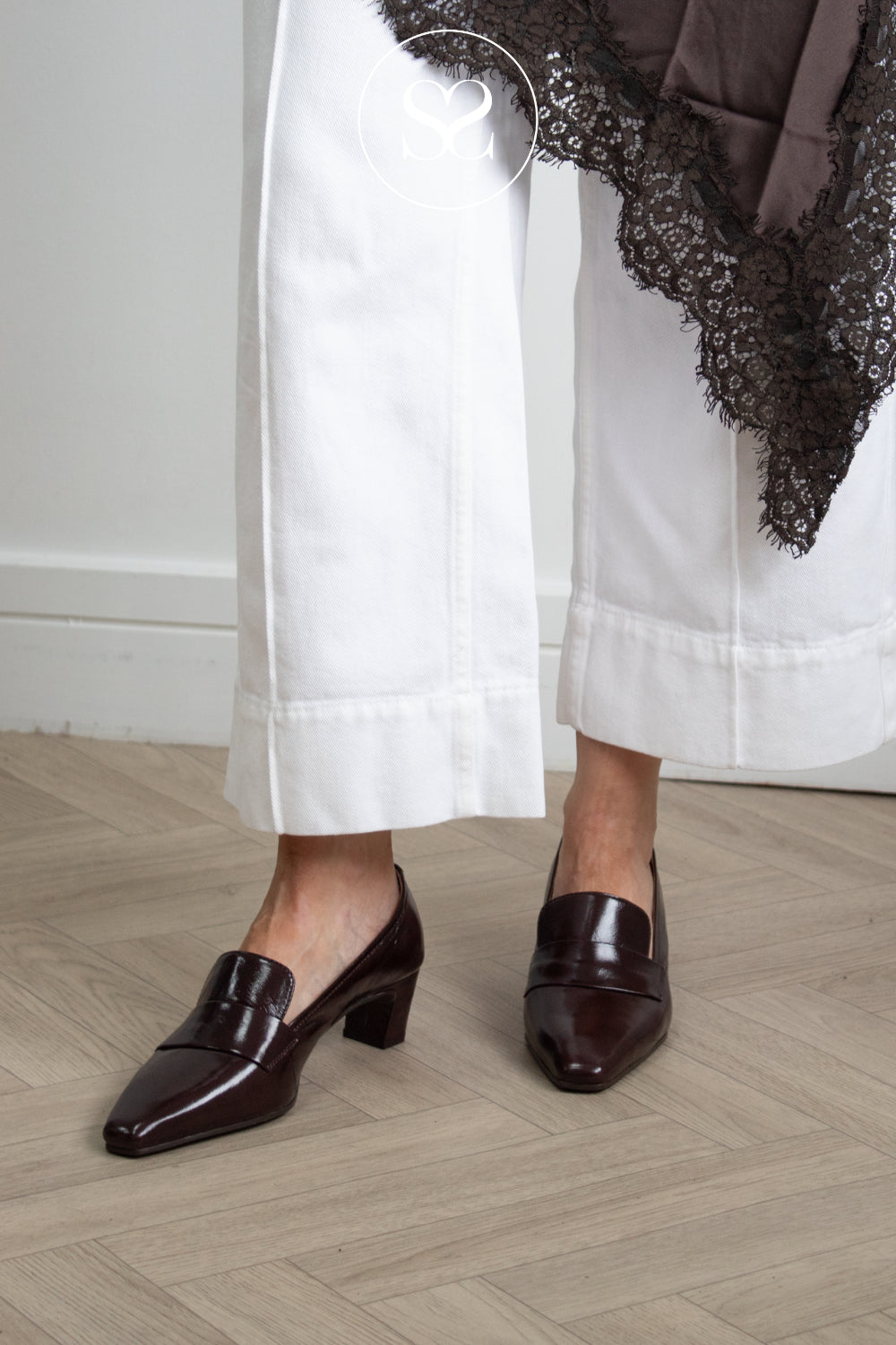 A person wearing dark brown high shine leather loafers with a low block heel and pointed toe, paired with white cropped trousers and a brown lace-trimmed top, standing on a light wood herringbone floor.
