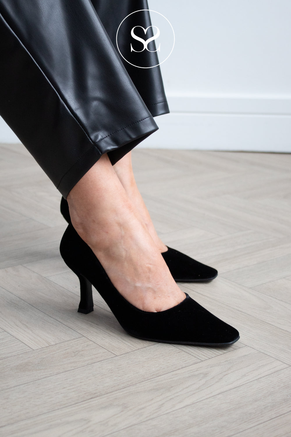 Lodi Pauline - Black velvet pointed-toe kitten heel pumps worn with black leather trousers, shown on a model standing on a light wood herringbone floor.