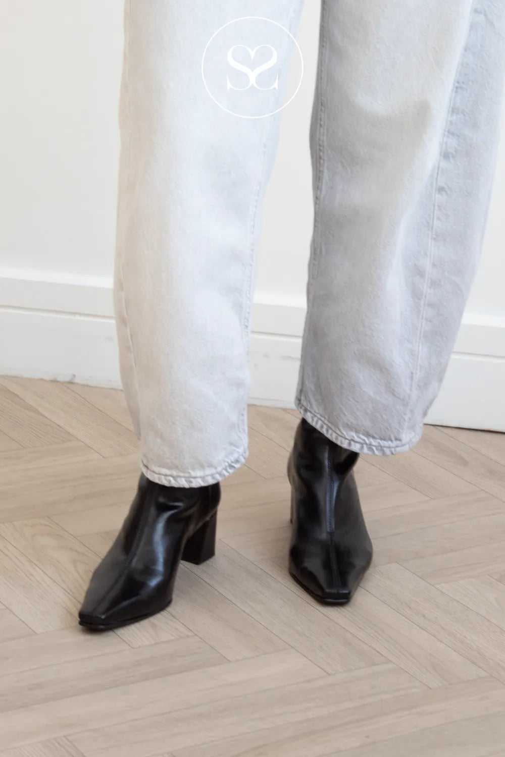A person wearing light-wash wide-leg jeans and Miss Elastic black pointed-toe ankle boots with block heels, standing on a light wood herringbone floor against a white wall.