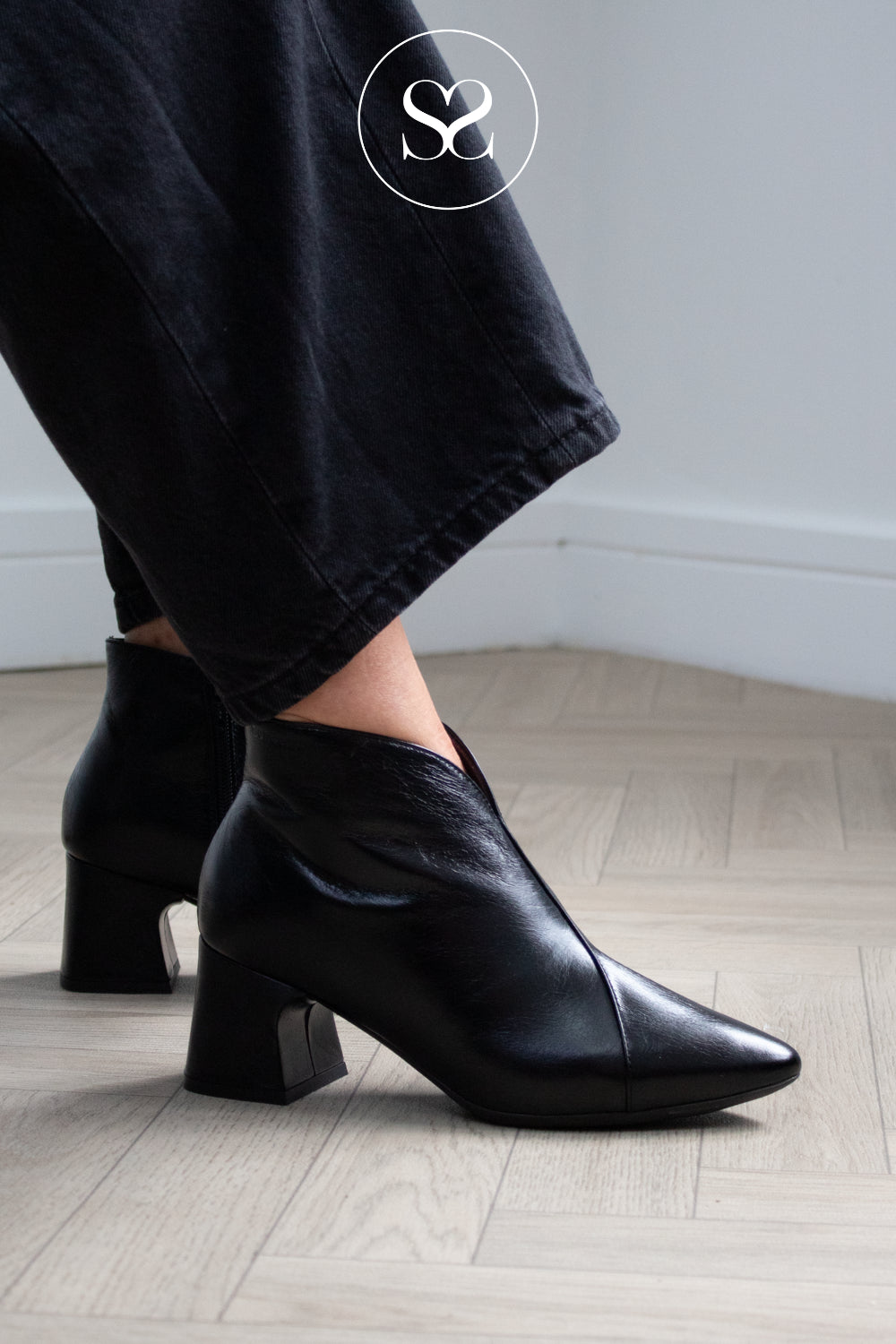 Black leather ankle boots with block heels and pointed toes, shown on a model wearing black jeans on a light wood herringbone floor. A white circular logo with 'Sheneil Shoes' is visible in the top right. In Ireland