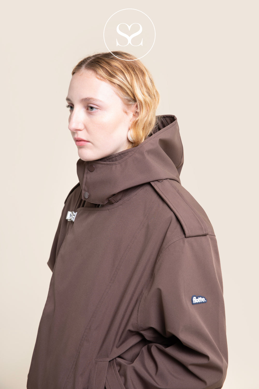 A model wearing a brown hooded jacket with a high collar, shown in profile against a solid beige background. The jacket features a small blue and white flotte logo on the left sleeve.