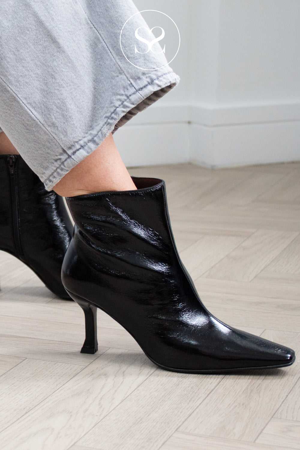A close-up of a person wearing glossy black pointed-toe ankle boots with kitten heels, paired with light grey cropped jeans, standing on a light wood floor. Lodi Boots in high shine leather