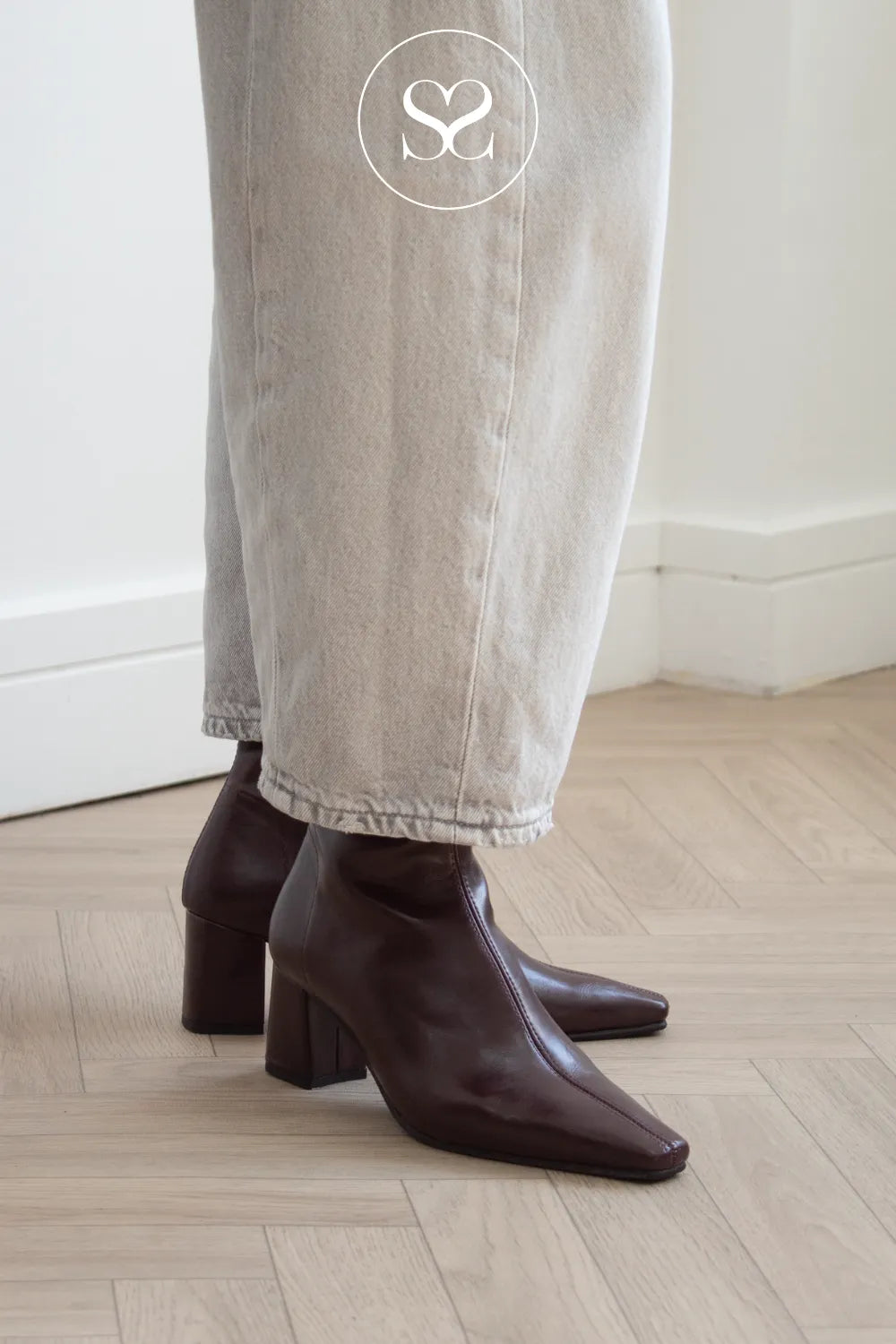 A person wearing dark burgundy pointed-toe ankle boots with block heels, paired with light beige wide-leg jeans, standing on a light wood herringbone floor.