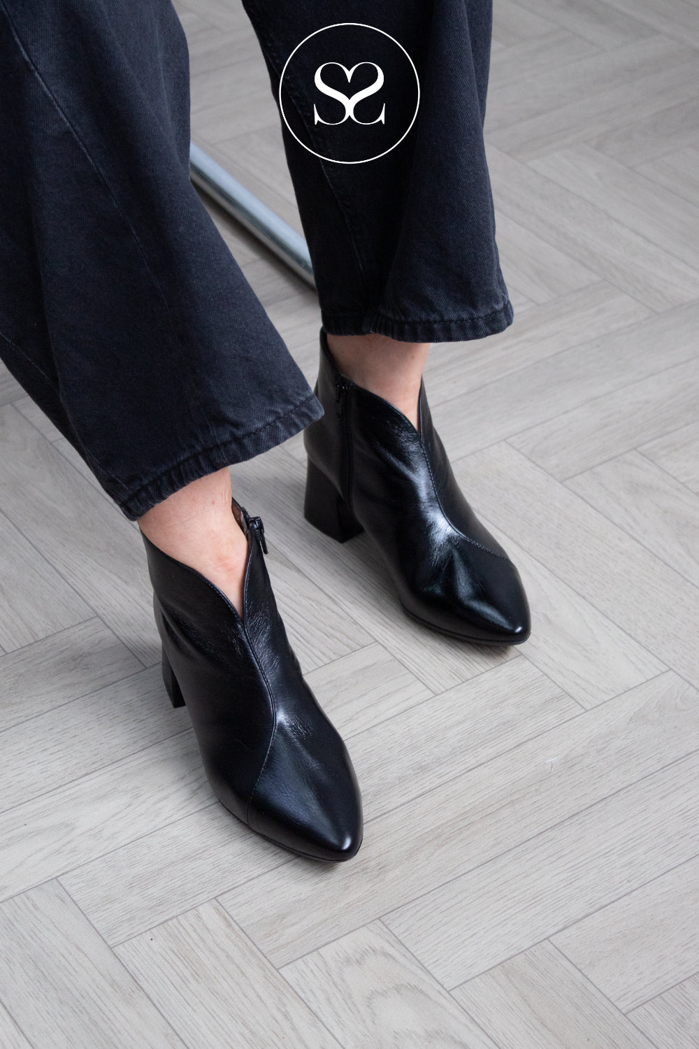 A person wearing Wonders black leather pointed-toe ankle boots with a low block heel and side zipper, paired with dark jeans, standing on a light gray wood plank floor.