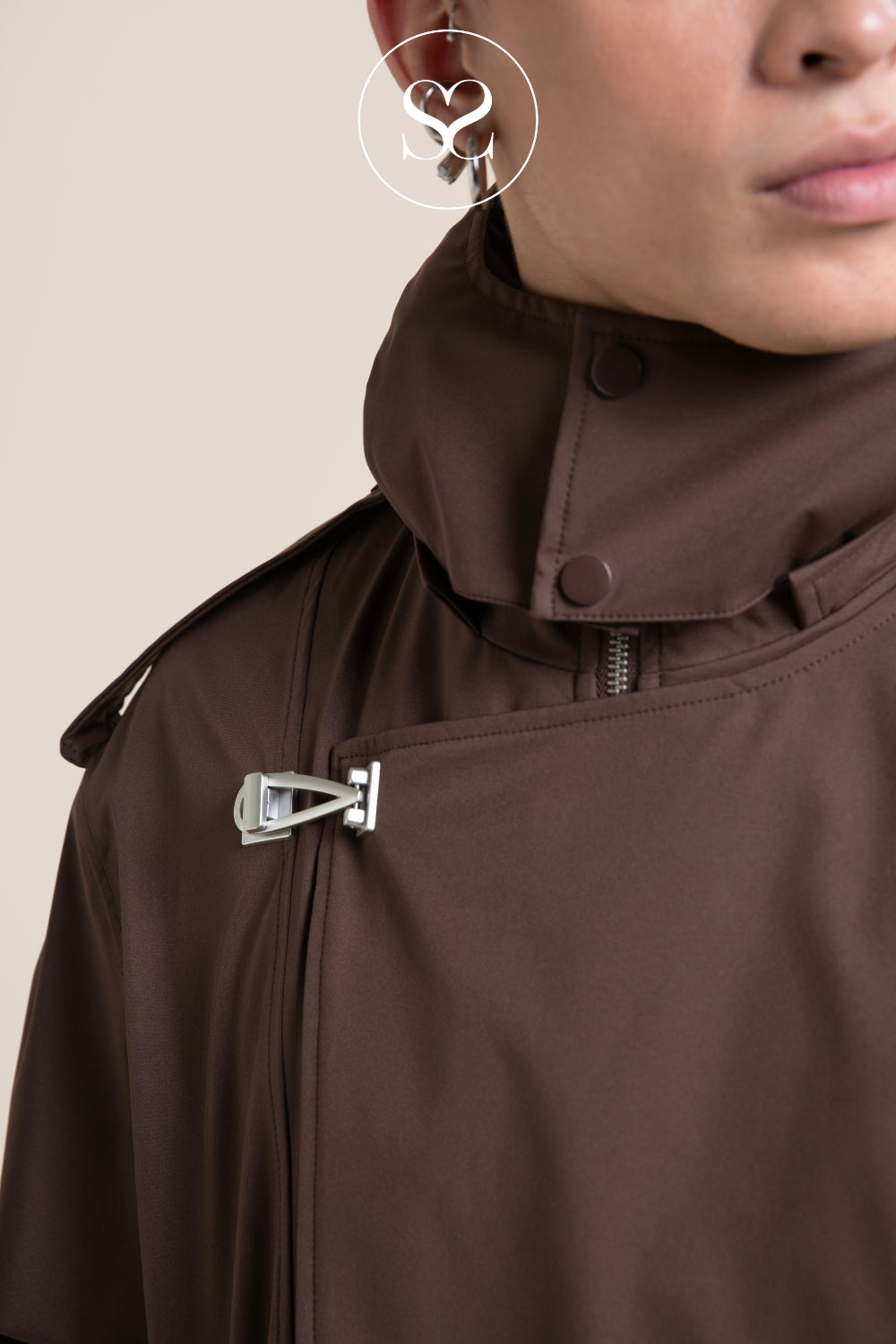 Close-up of a person wearing a dark brown high-collar jacket with a silver metal toggle closure and a visible zipper, against a plain beige background.