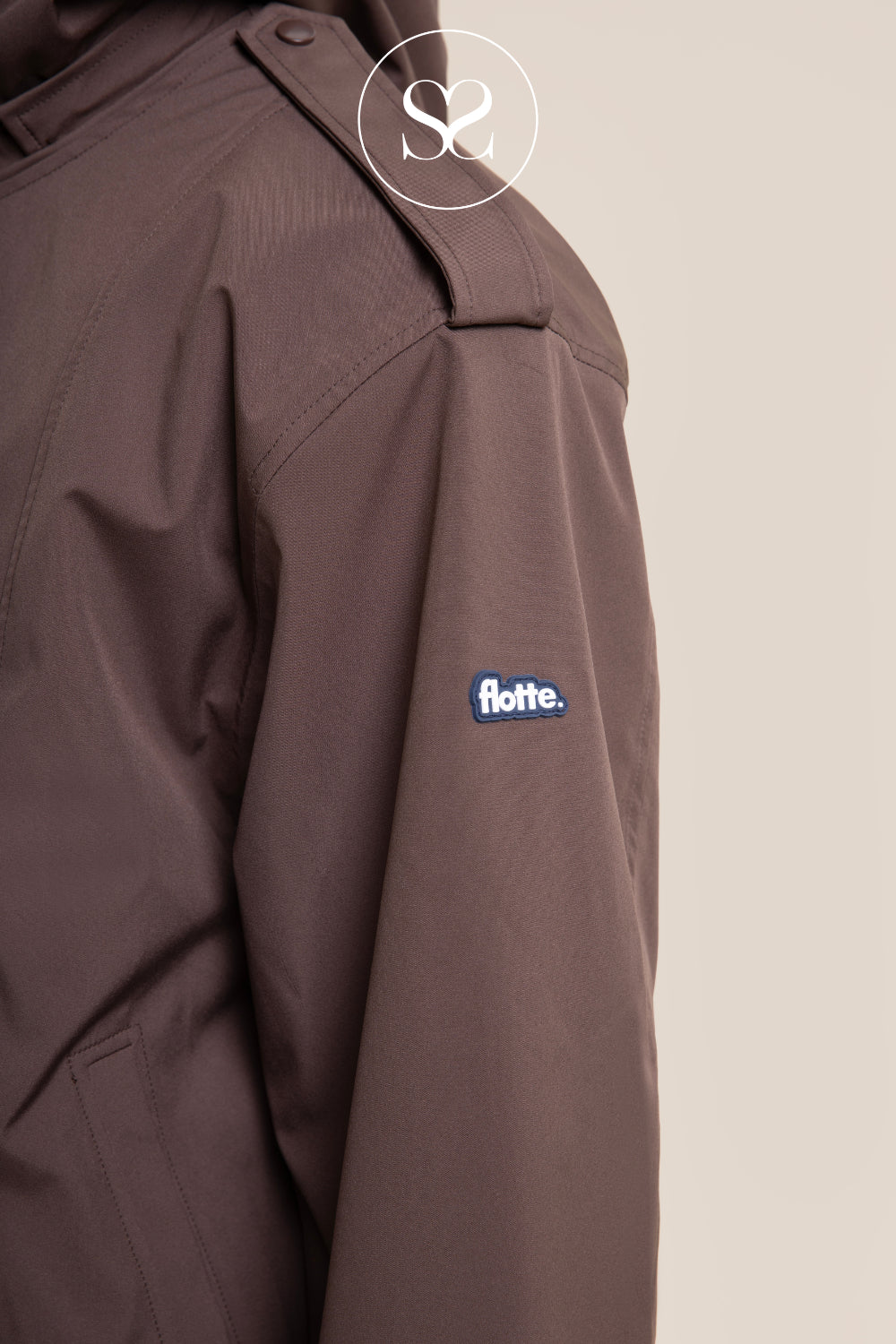 Close-up of a dark brown jacket sleeve featuring a blue and white 'flotte.' logo patch on the upper arm, set against a solid light beige background.