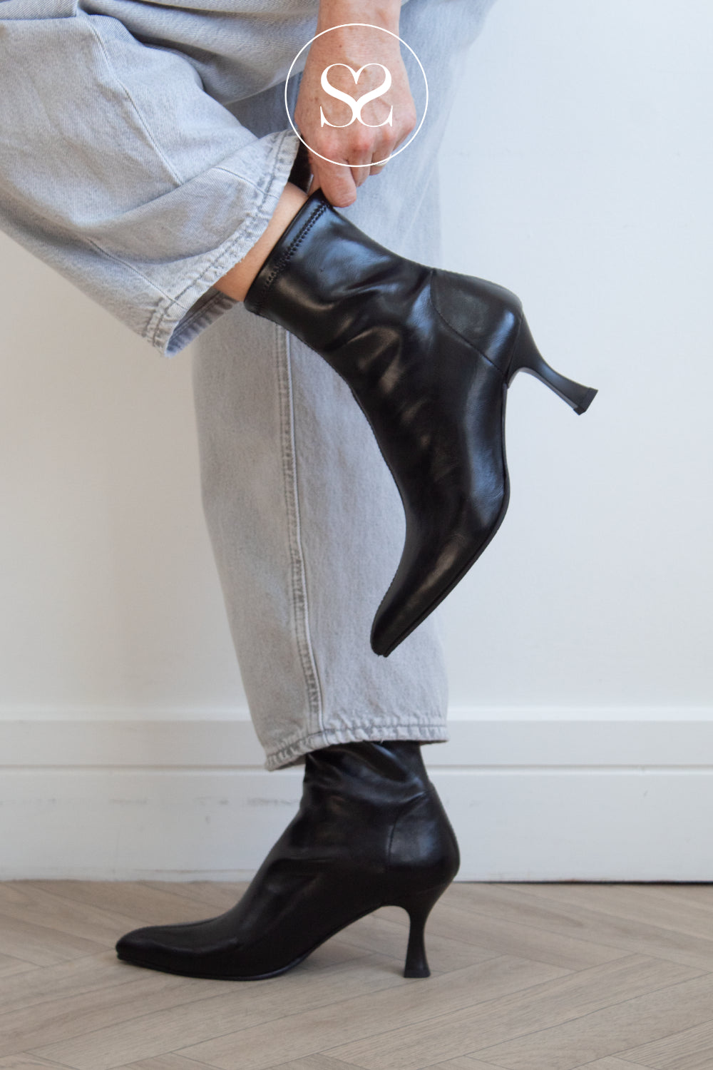 A person wearing black pointed-toe sock boots with kitten heels from Miss Elastic, paired with light-wash denim jeans, shown from the waist down against a white wall and light wood floor.