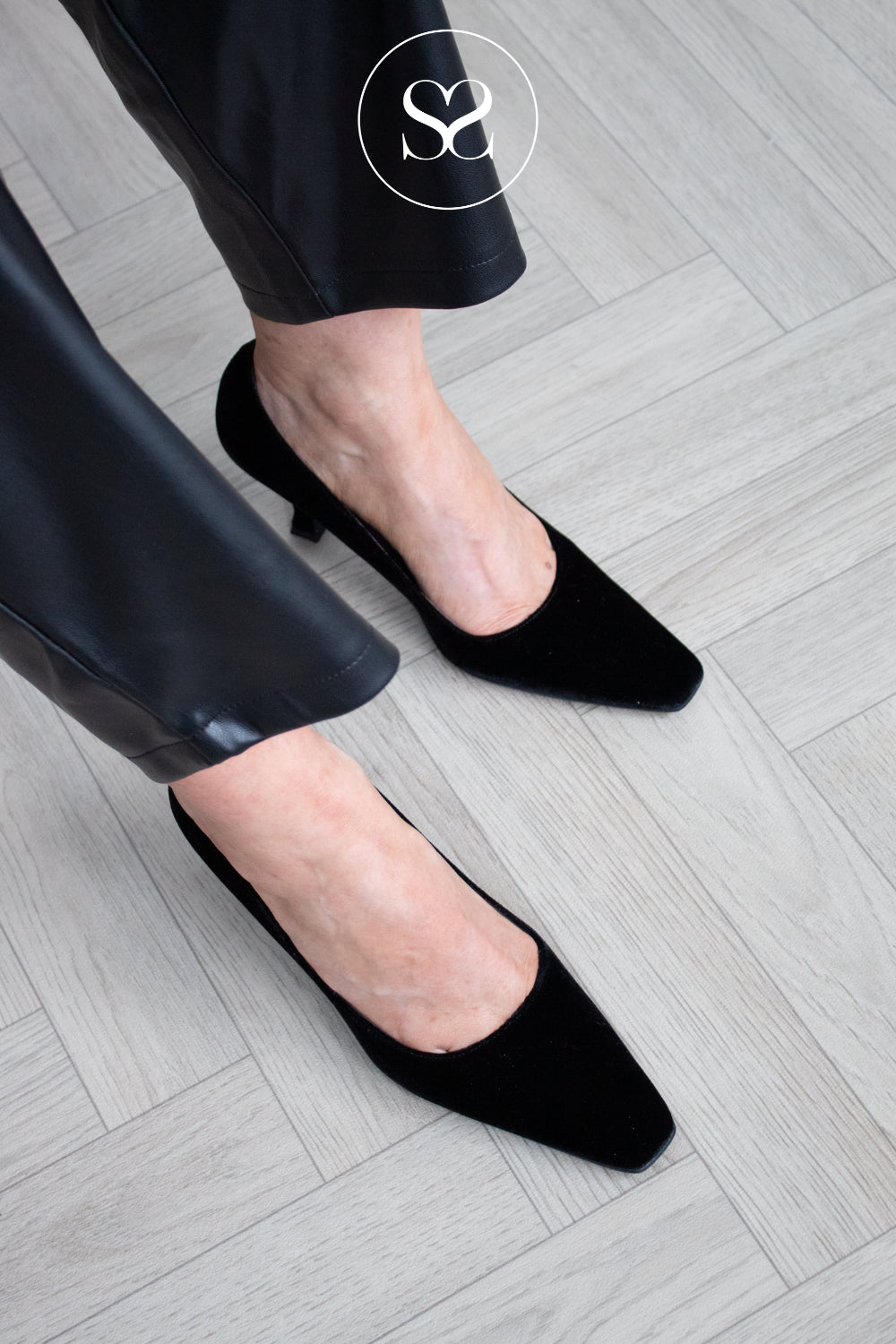 A close-up lifestyle shot of a person wearing black velvet pointed-toe kitten heel pumps with black pants, standing on a light gray wood herringbone floor. A circular 'SS' Sheneil Shoes logo watermark is visible at the top. Shoes are lodi - available in Ireland