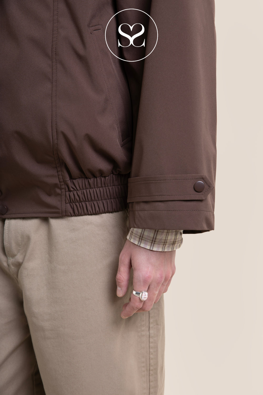 Close-up of a woman wearing a brown jacket with a buttoned cuff and elastic hem, paired with beige trousers and a plaid shirt cuff, against a light beige background.