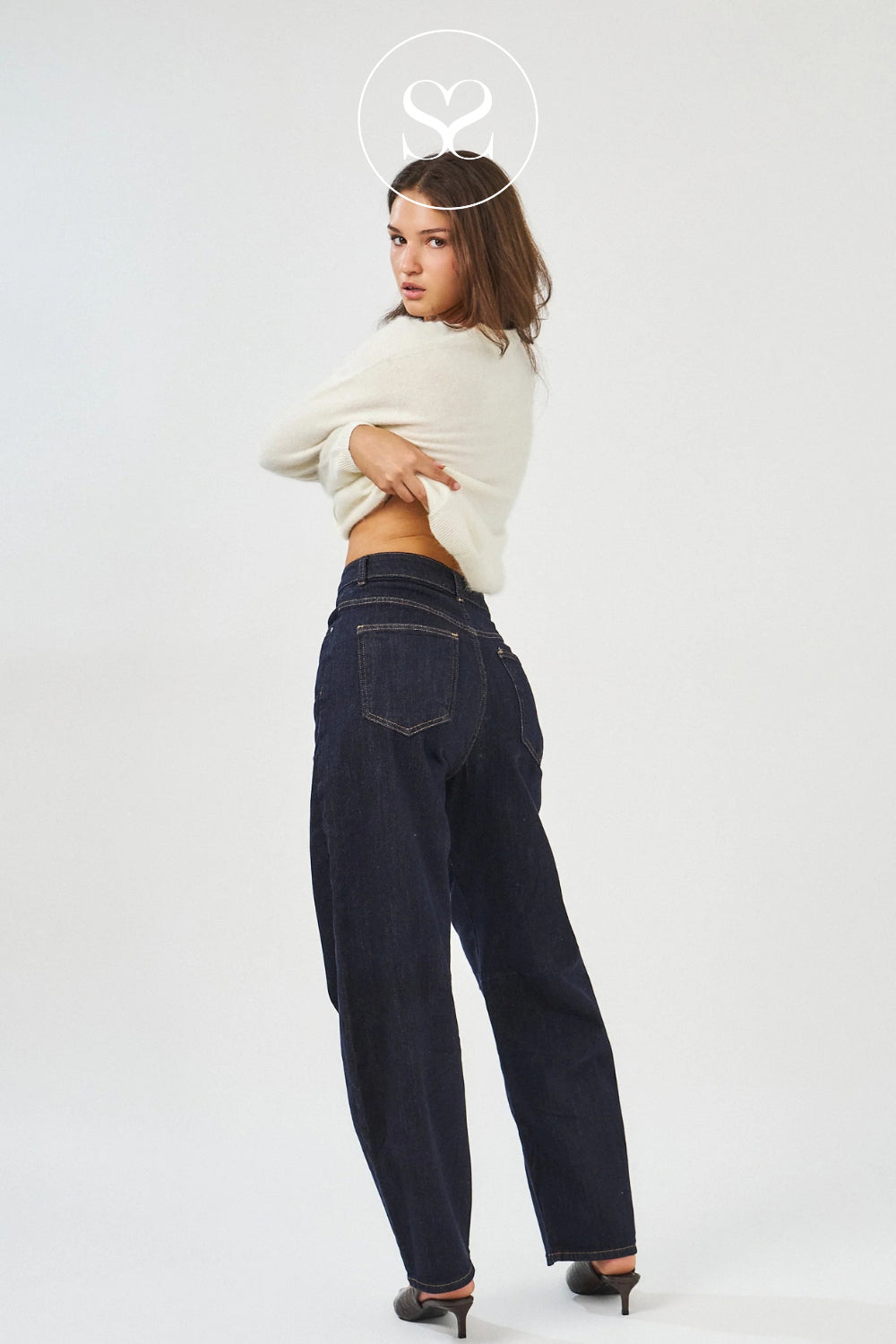 A woman posing from the back wearing dark wash wide-leg jeans, a cream ribbed knit long-sleeve crop top, and black pointed-toe heels against a plain white studio background.
