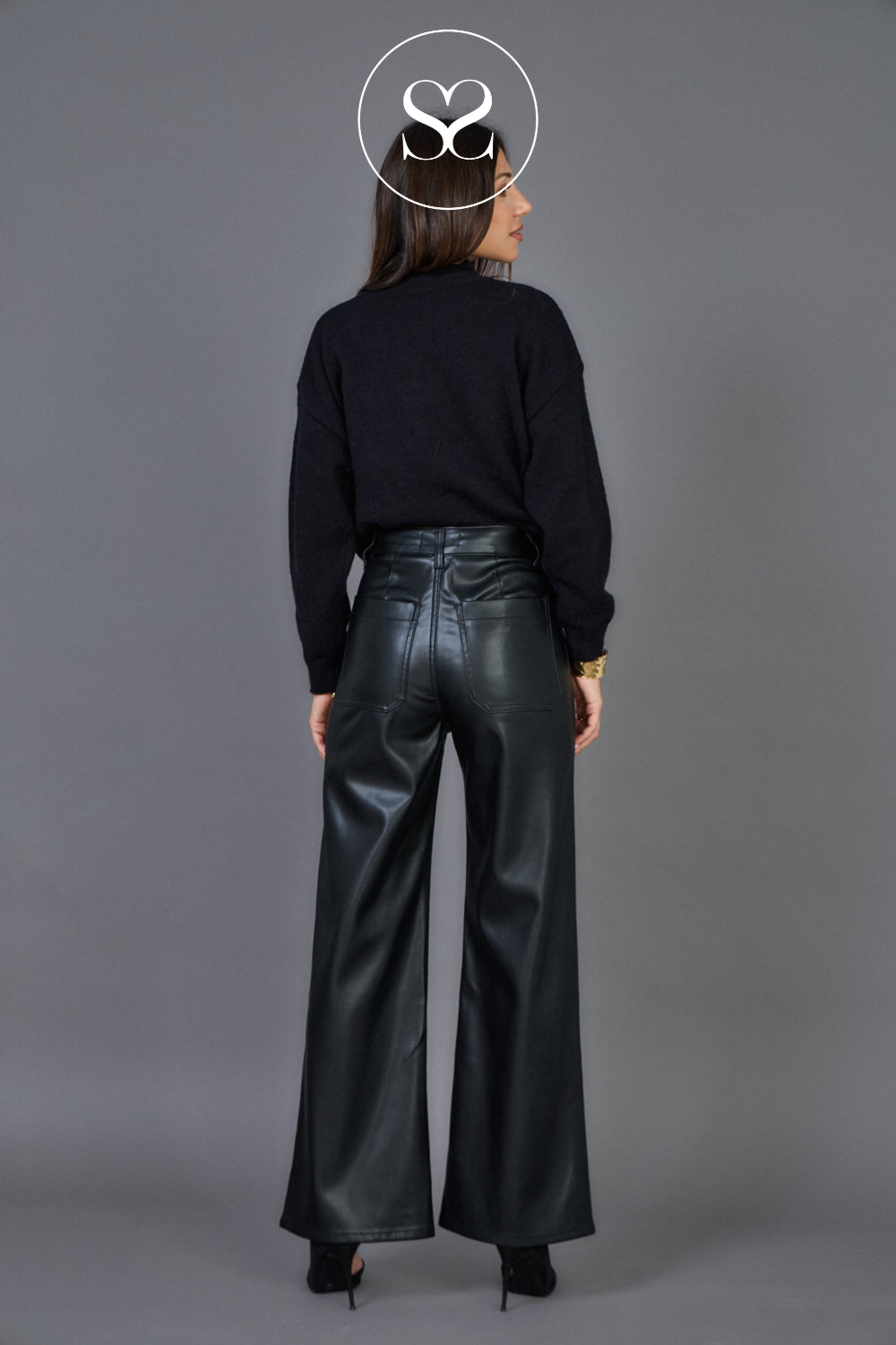 A woman viewed from the back wearing a black long-sleeve top and shiny black wide-leg pants, standing against a plain gray studio background. 
