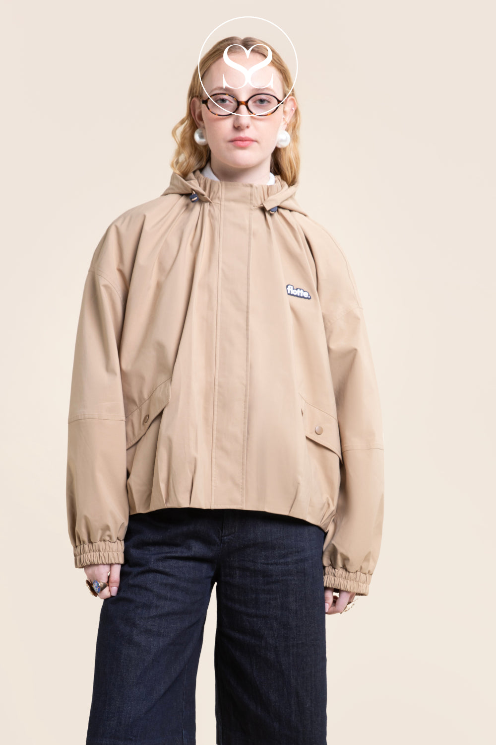 A model wearing a beige hooded jacket from flotte with a small embroidered logo on the chest, paired with dark blue jeans, standing against a solid light beige background.