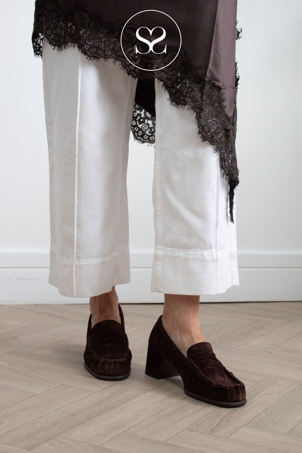 Studio Image: A woman wearing  dark brown suede tassel loafers with low block heel from unisa, standing on a light wood floor against a white wall. A sheer taupe fabric with black lace is draped over the upper leg, and a circular 'SS' logo watermark is visible near the top.