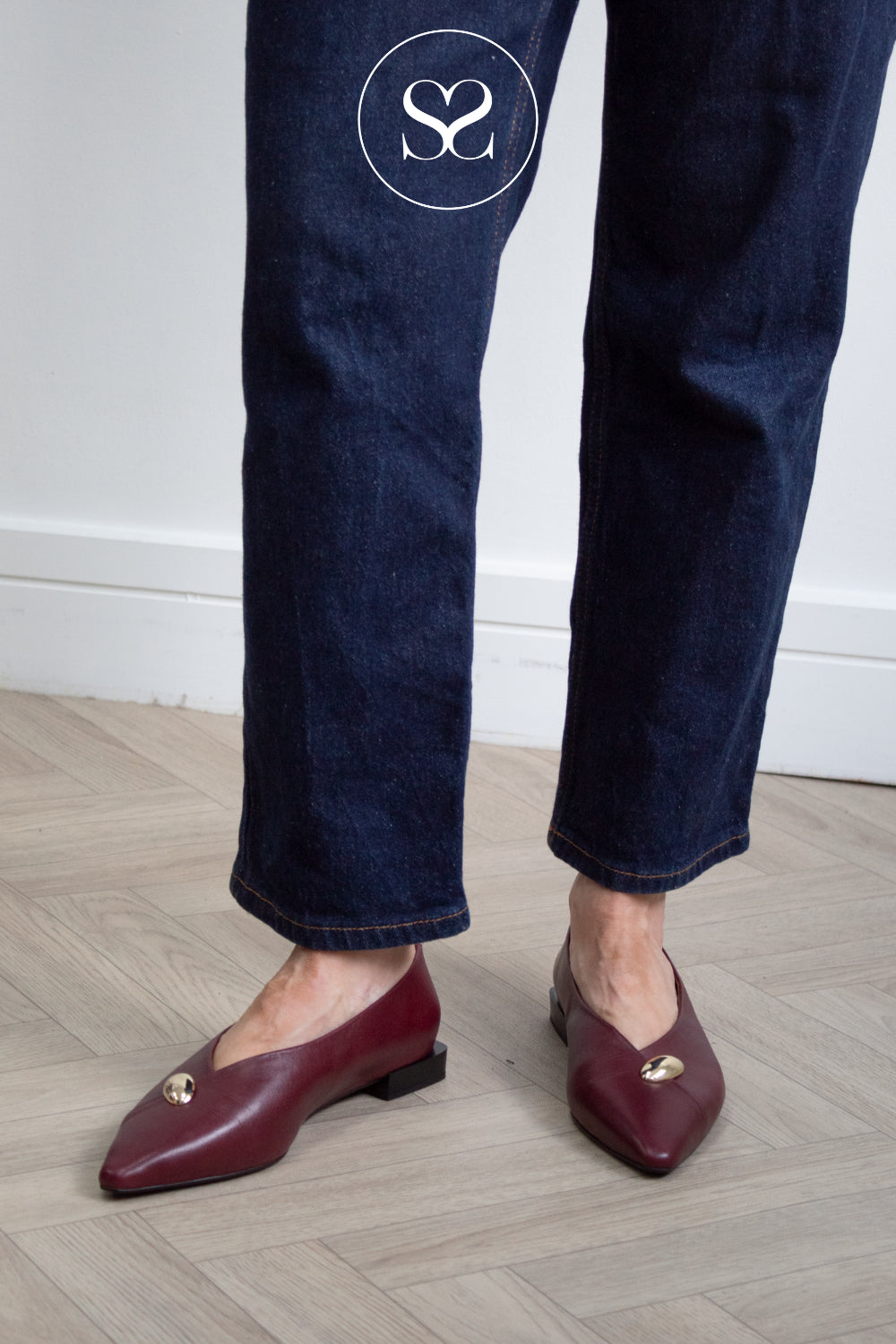 A person wearing burgundy leather  pointed-toe flats with small gold hardware, standing on a light wood herringbone floor against a white wall.