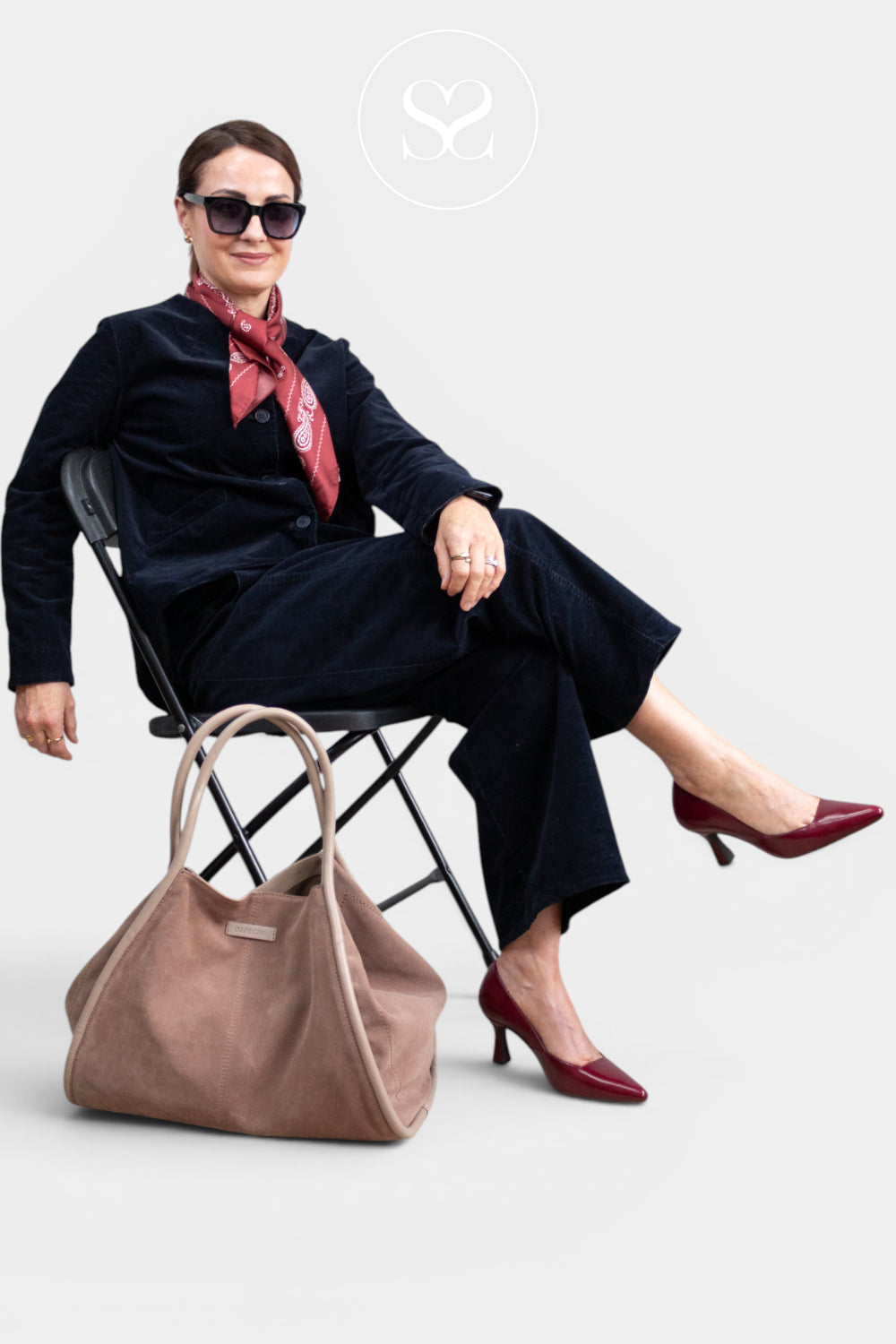 Editorial Image: A woman sits on a folding chair wearing a dark navy velvet suit, a red patterned scarf, sunglasses, and burgundy heels, with a large Depeche  taupe-brown suede tote bag on the floor beside her. A circular 'Sheneil Shoes' logo is visible at the top center.