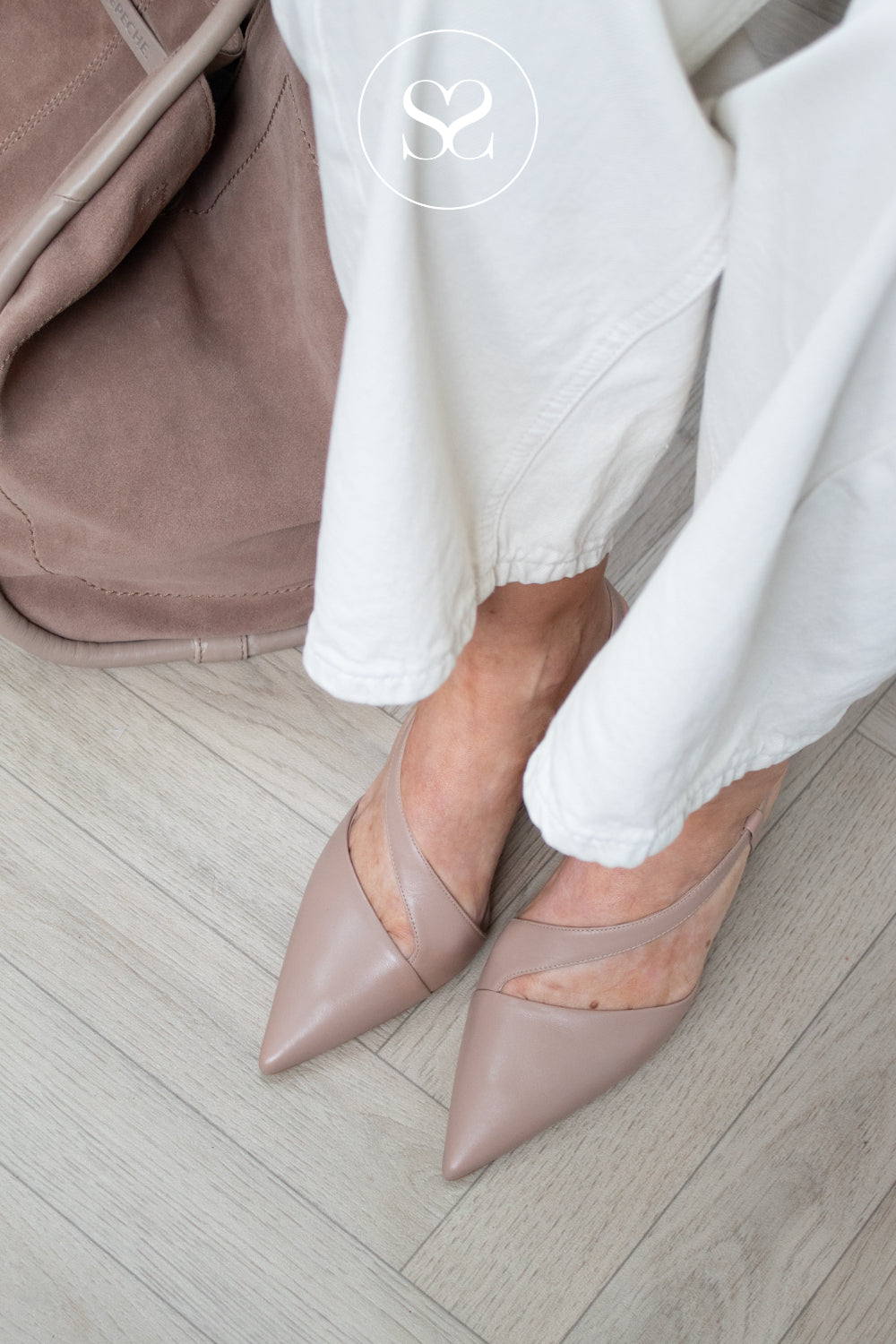 A close-up lifestyle shot of a person wearing lodi taupe pointed-toe slingback heels with white pants, standing on a light wood floor next to a matching Depeche taupe bag.