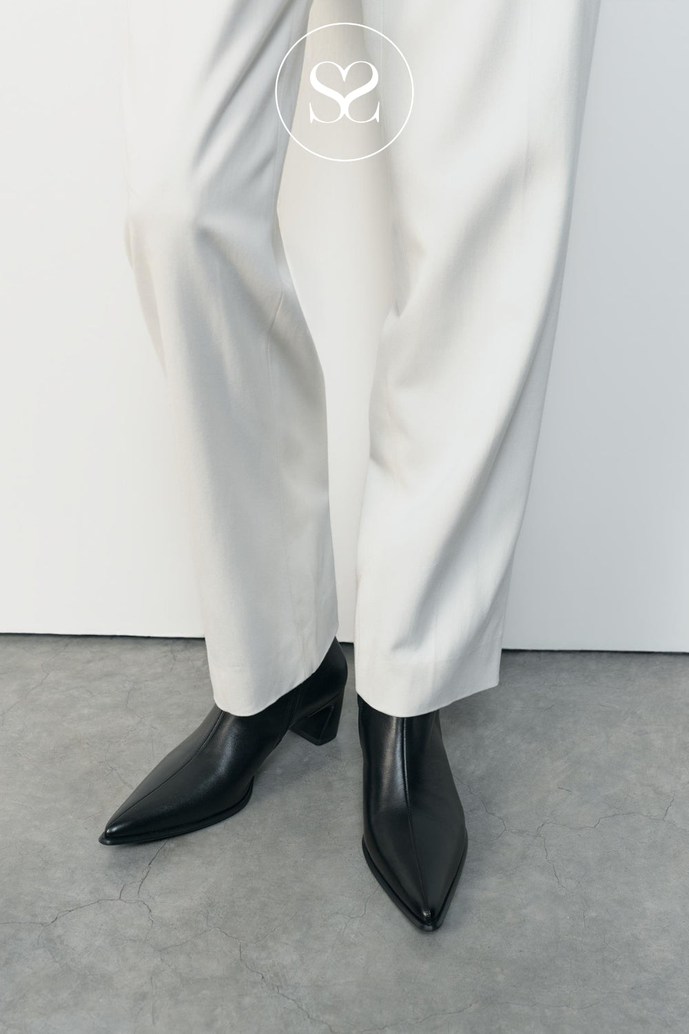 A close-up of a women's legs wearing black pointed-toe ankle boots with low block heels from Vagabond and white wide-leg trousers, standing on a gray concrete floor against a white wall.