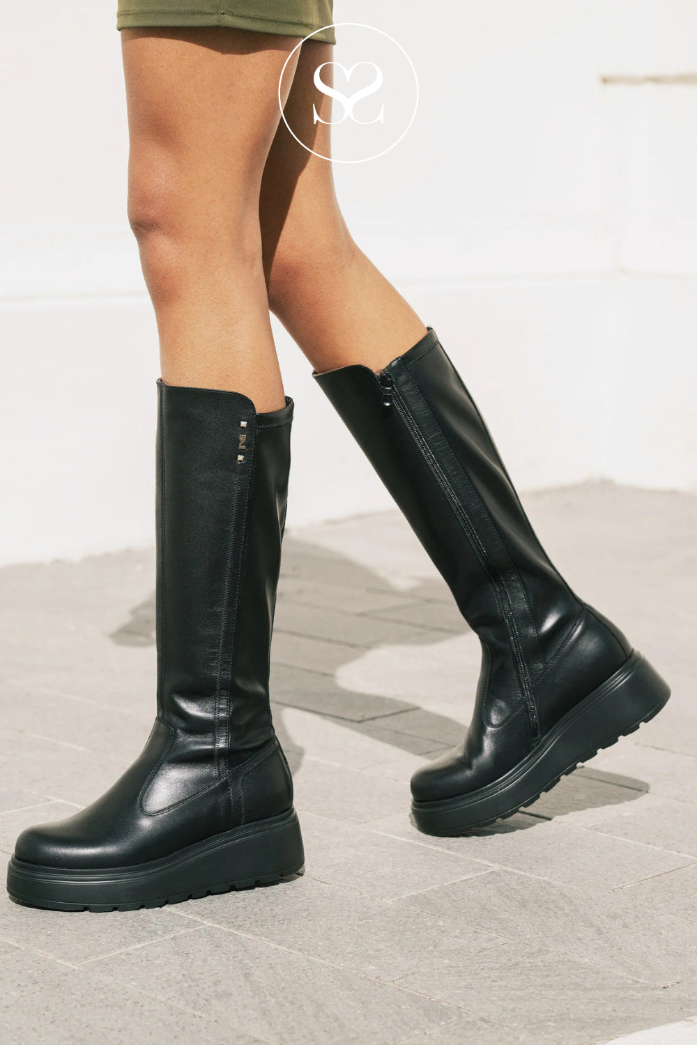 A woman wearing a pair of Nero Giardini black knee-high platform boots with side zippers, standing on a light gray tiled floor against a white wall.