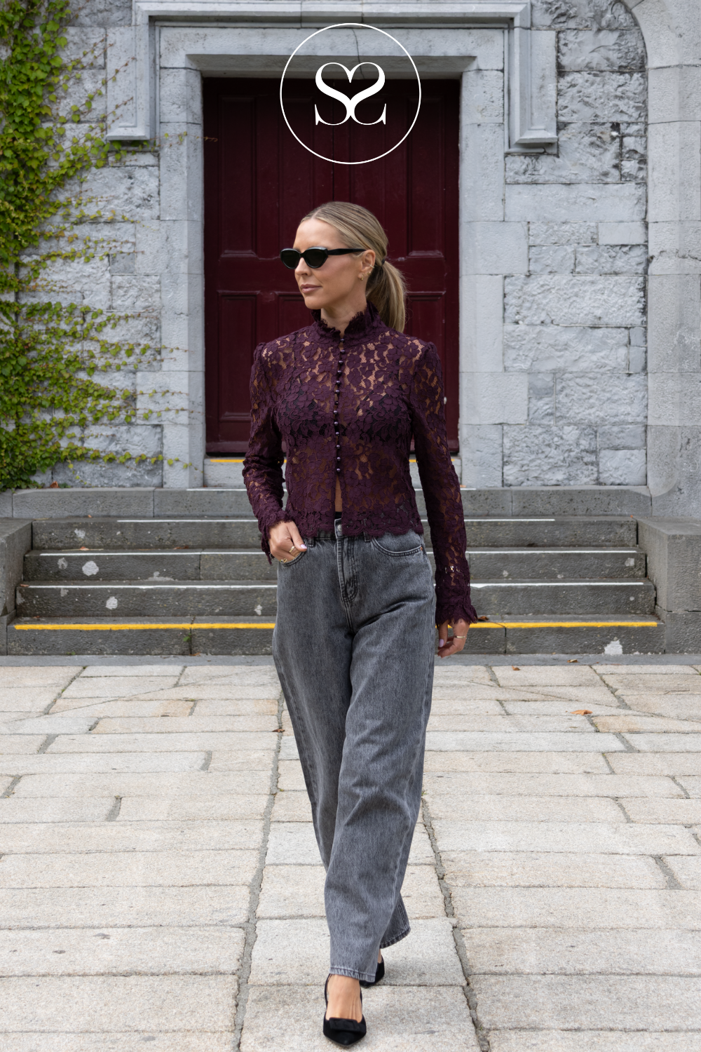 A woman walking outdoors in front of a stone building with a dark red door, wearing a dark burgundy lace long-sleeve top, wide-leg gray jeans, and black heels. A circular 'S2' logo is visible at the top center.