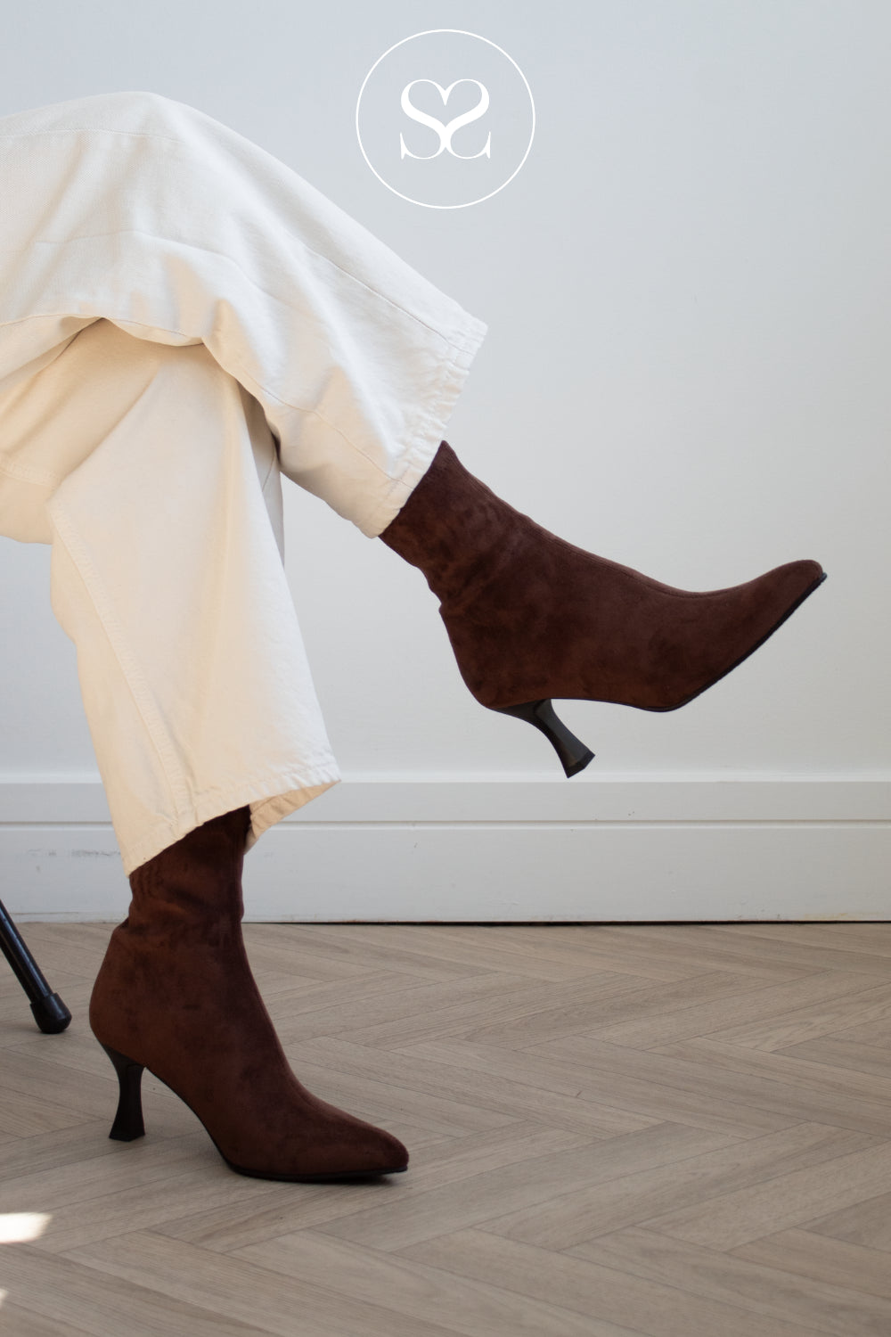 A person wearing dark brown suede ankle boots with kitten heels and cream-colored wide-leg trousers, sitting on a chair in a room with a light gray wall and herringbone wood floor. A circular logo with an 'S' is visible at the top.