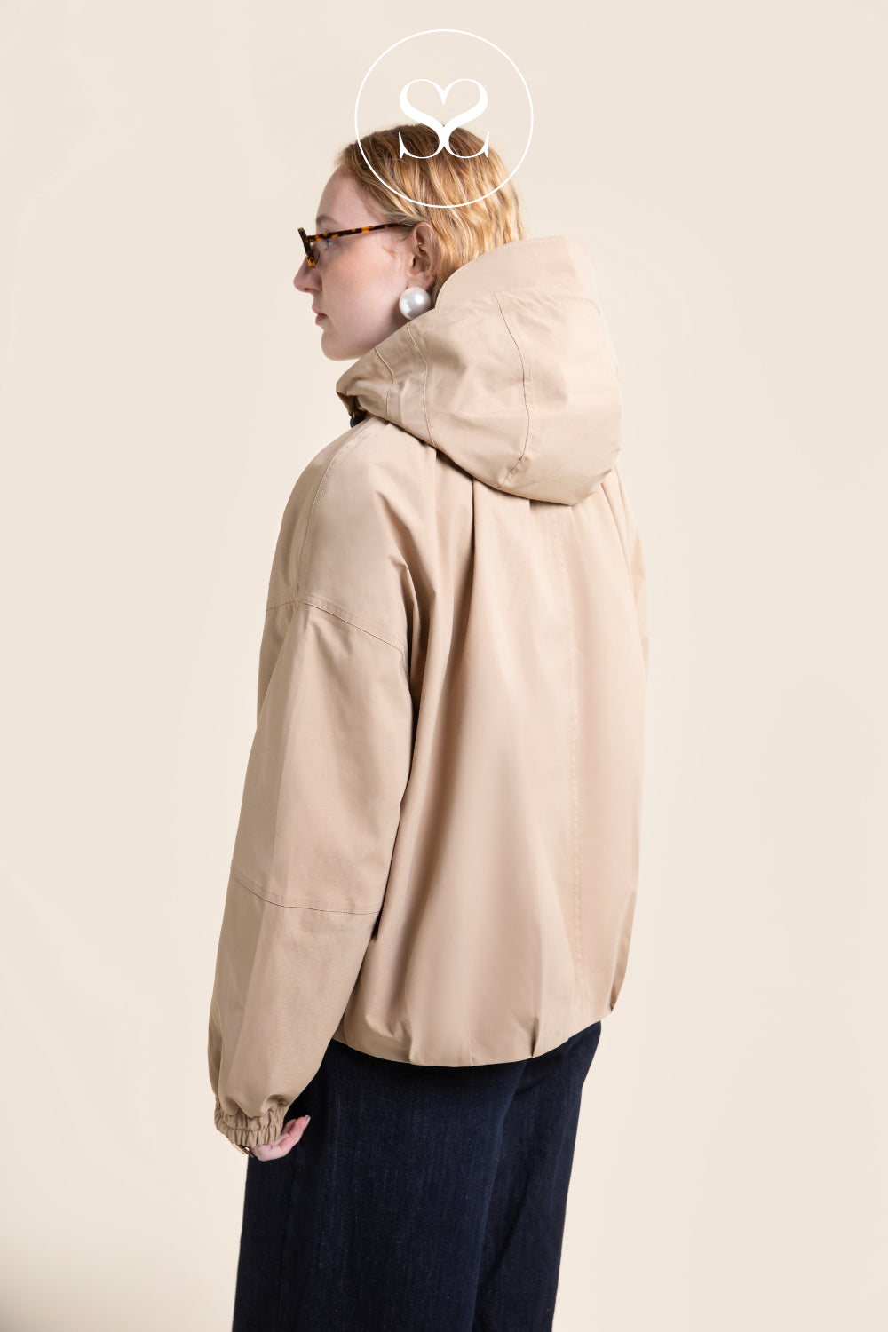 A woman shown from the back wearing a light beige flotte hooded jacket and dark navy pants, photographed against a plain warm beige studio background.