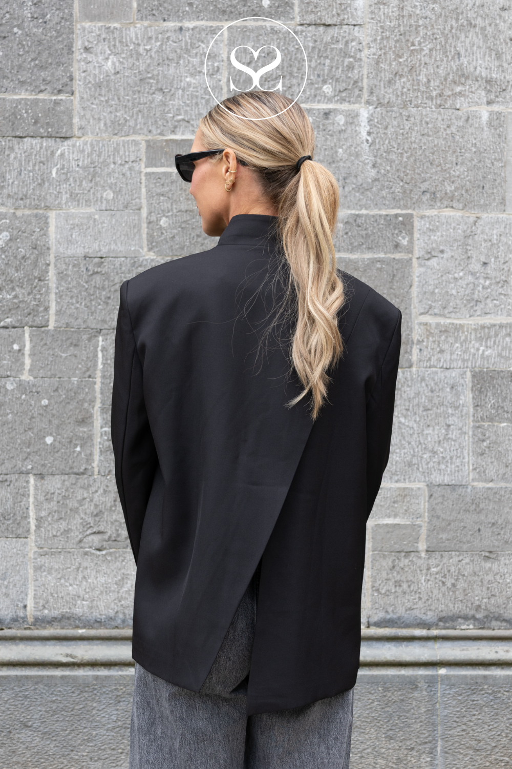 A woman with blonde hair in a ponytail, wearing a black asymmetrical wrap-style blazer and gray trousers, photographed from behind against a gray stone wall.