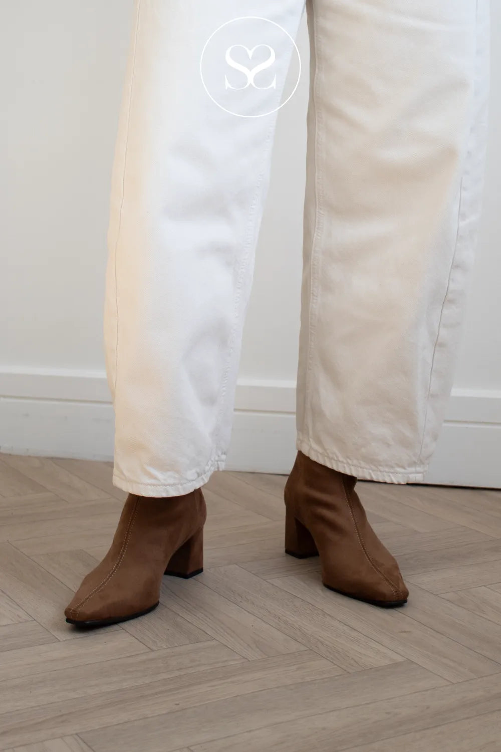A person wearing white wide-leg pants and brown ankle boots with a block heel, standing on a light wood herringbone floor against a white wall.